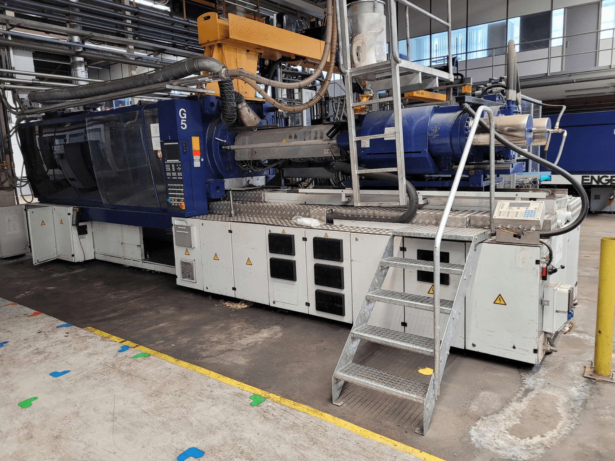Máquina de moldeo por inyección G5, vista lateral, con panel de control, escalera y superficie de trabajo de acero inoxidable visibles.