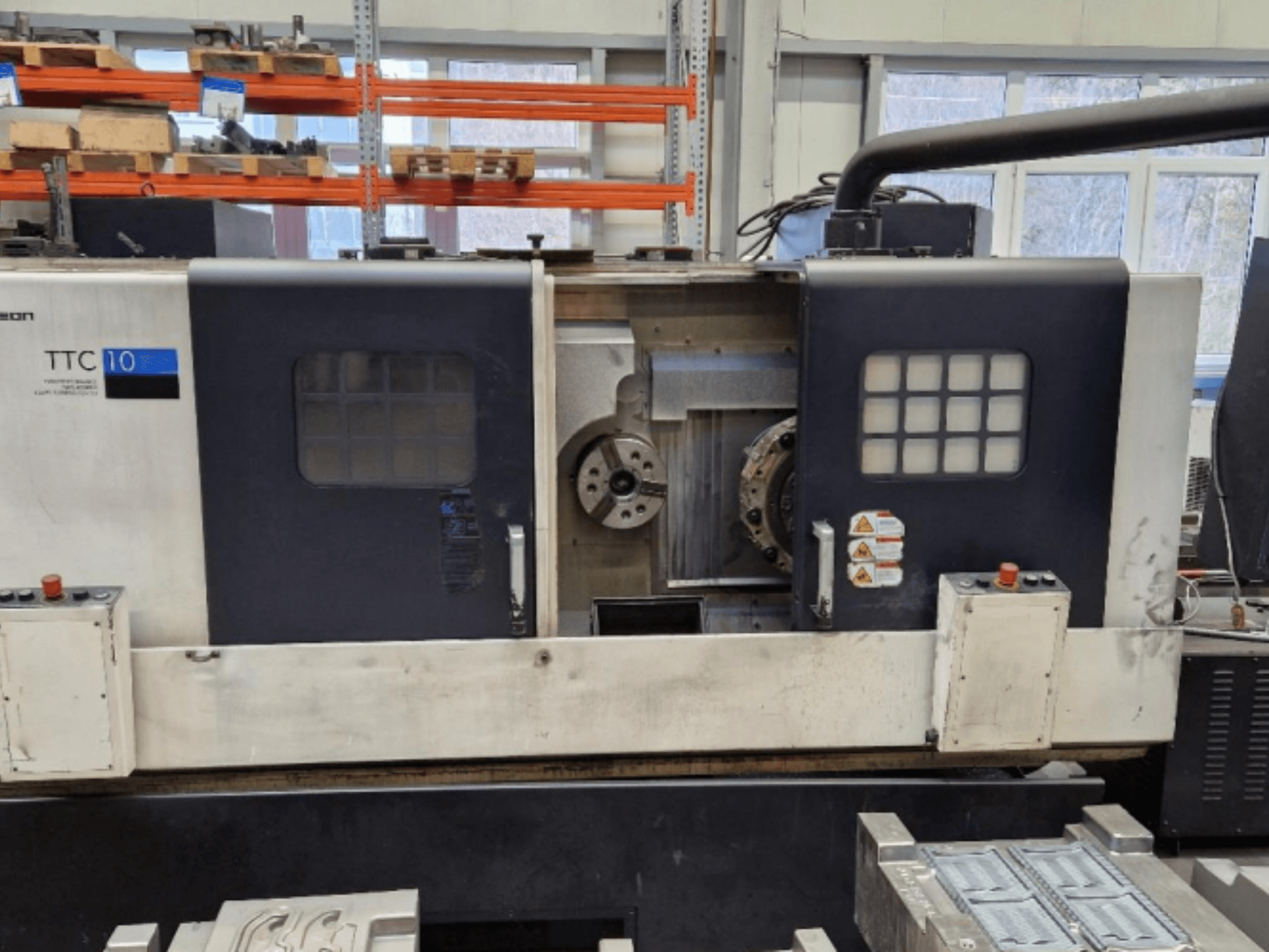 Vista frontal de la máquina de torno TTC 10, mostrando el panel de control, los mandriles visibles y el espacio de trabajo con piezas metálicas.