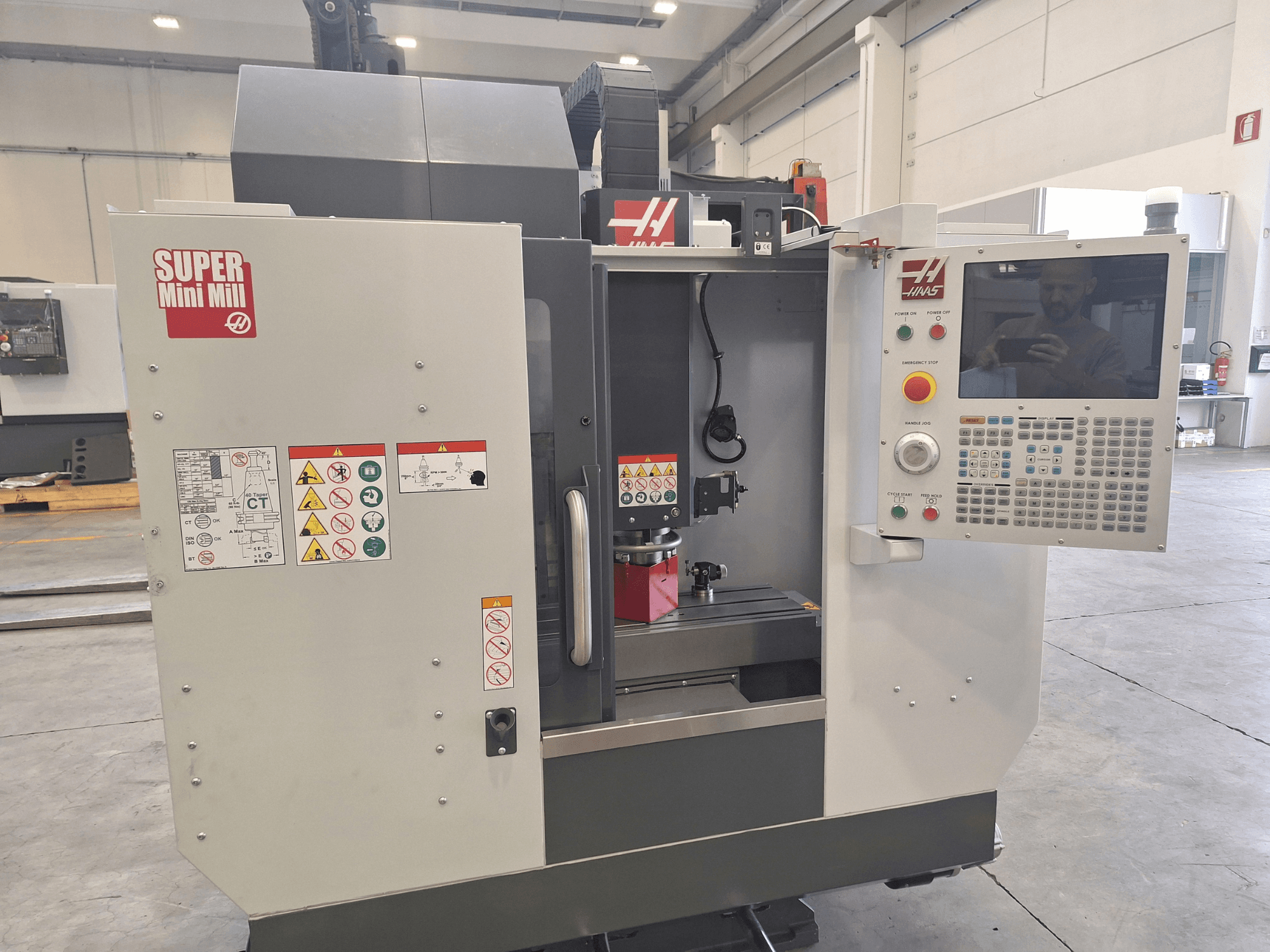 Vista frontal de la Super Mini Mill de HAAS, en la que se aprecian el panel de control, las etiquetas de seguridad y la zona de mecanizado con una pieza de trabajo montada.