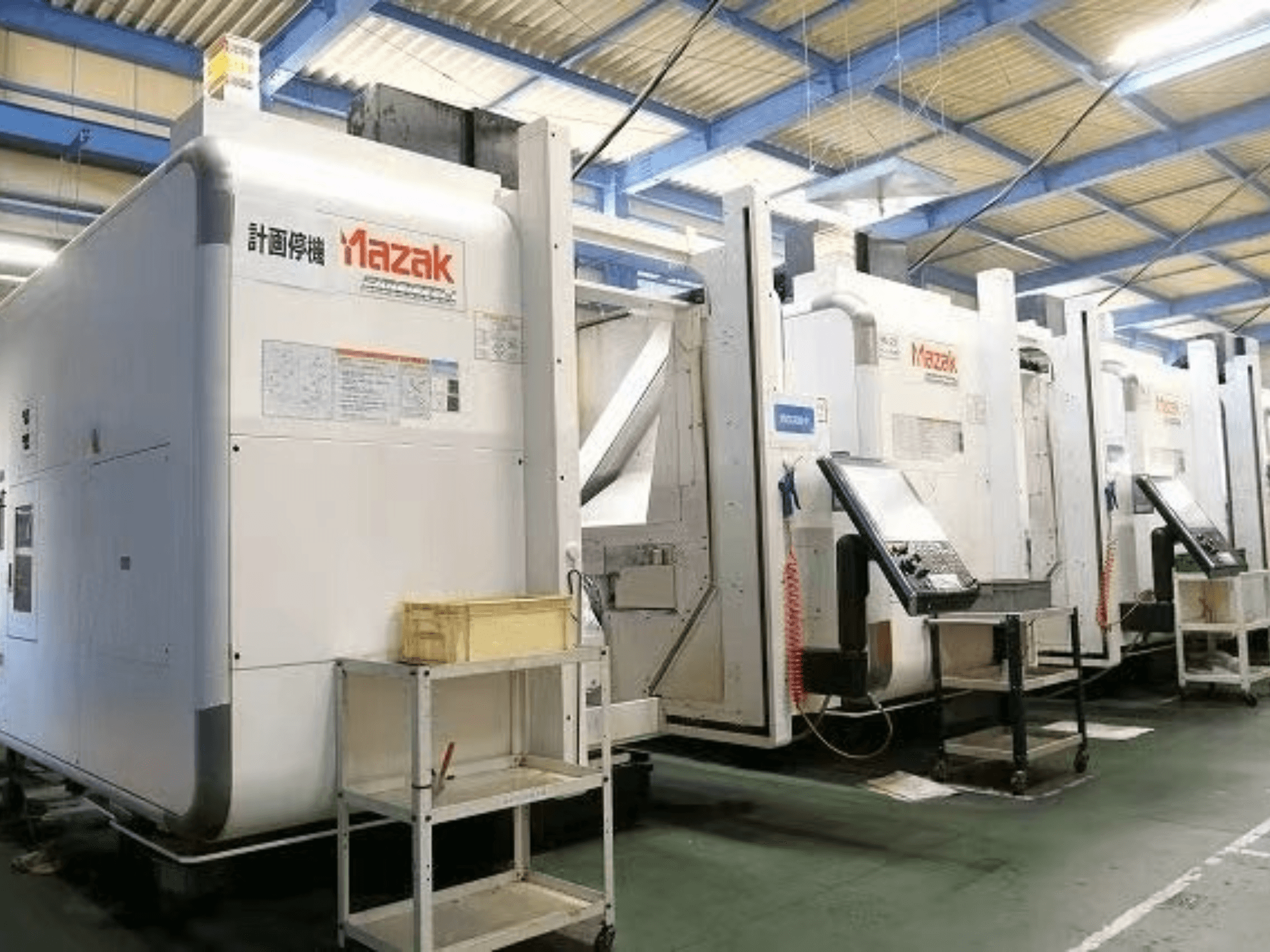 Máquinas CNC Mazak en un entorno de fábrica, vista lateral mostrando paneles de control y etiquetas sobre cuerpos blancos.