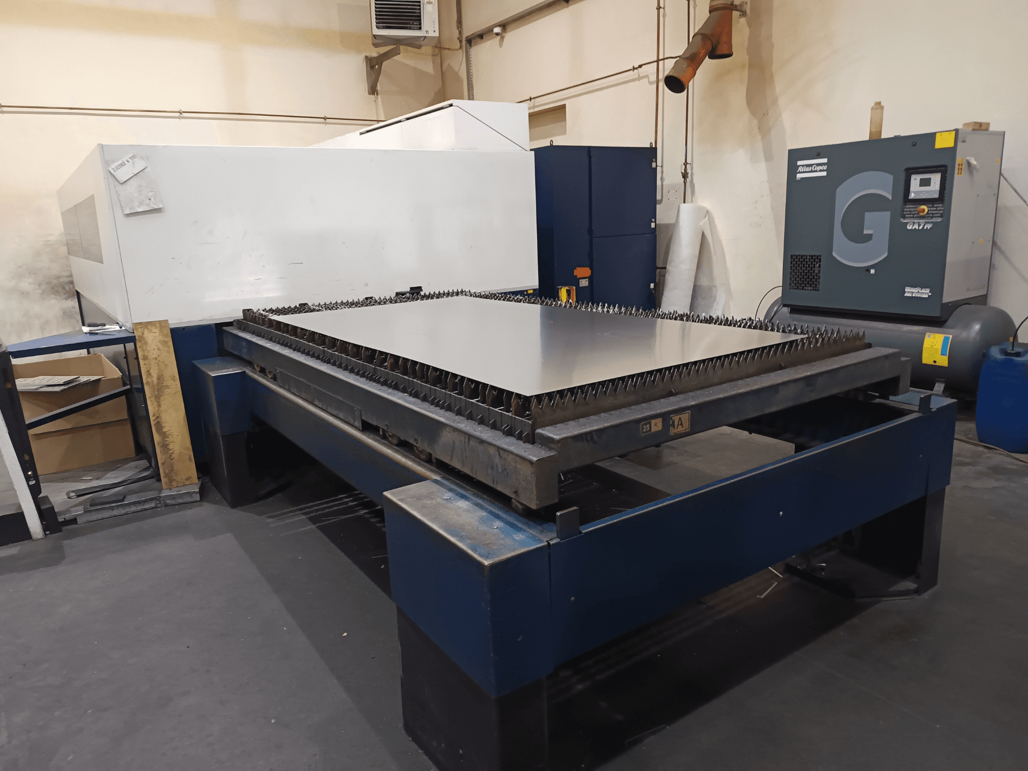 Máquina de corte industrial con mesa metálica, vista frontal. Presenta bordes afilados, compresor de aire a la derecha y bastidor de acero.