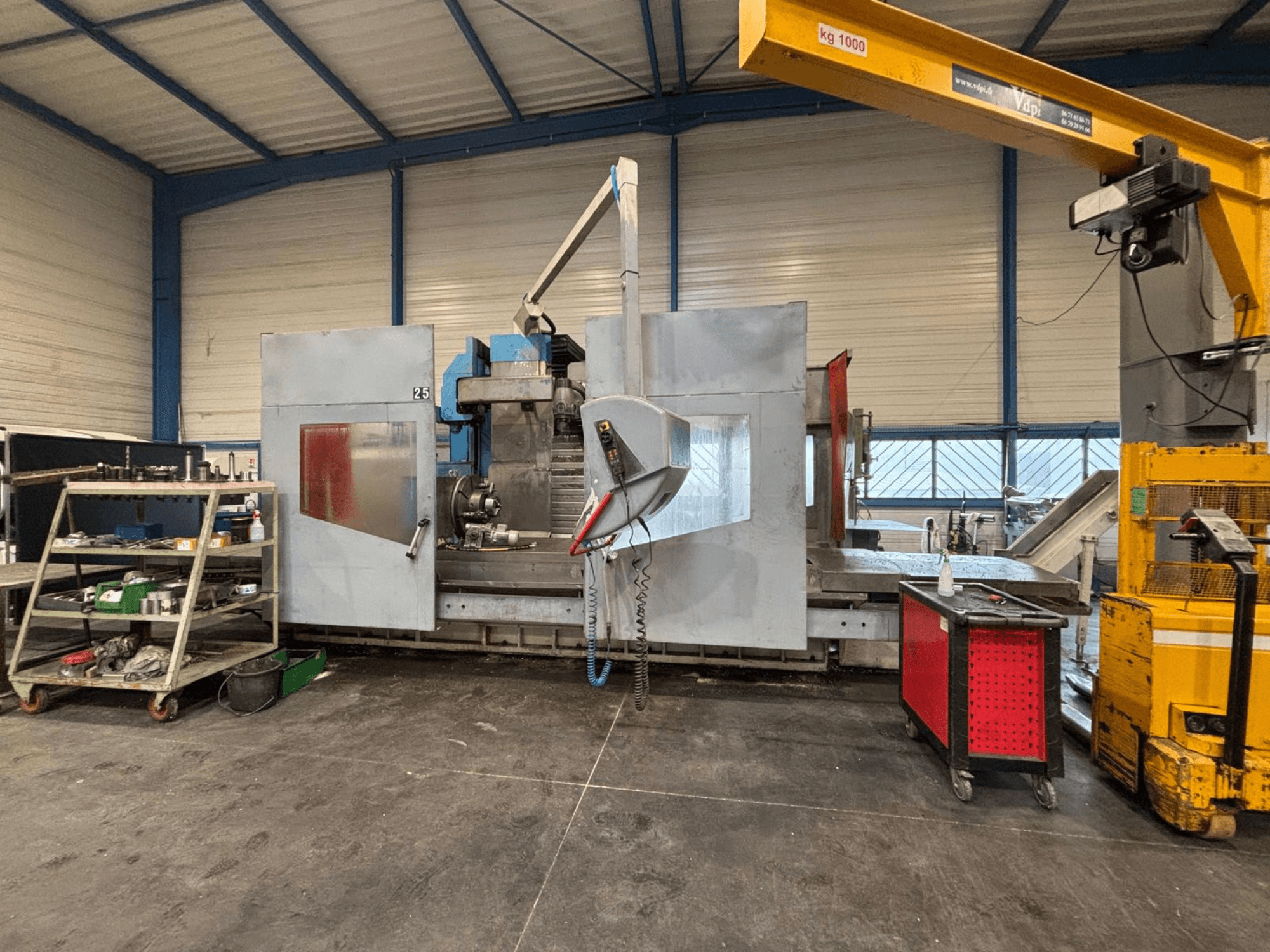 Fresadora CNC vista lateral en un taller; herramientas y estantes de almacenamiento visibles. Marca y modelo no especificados.