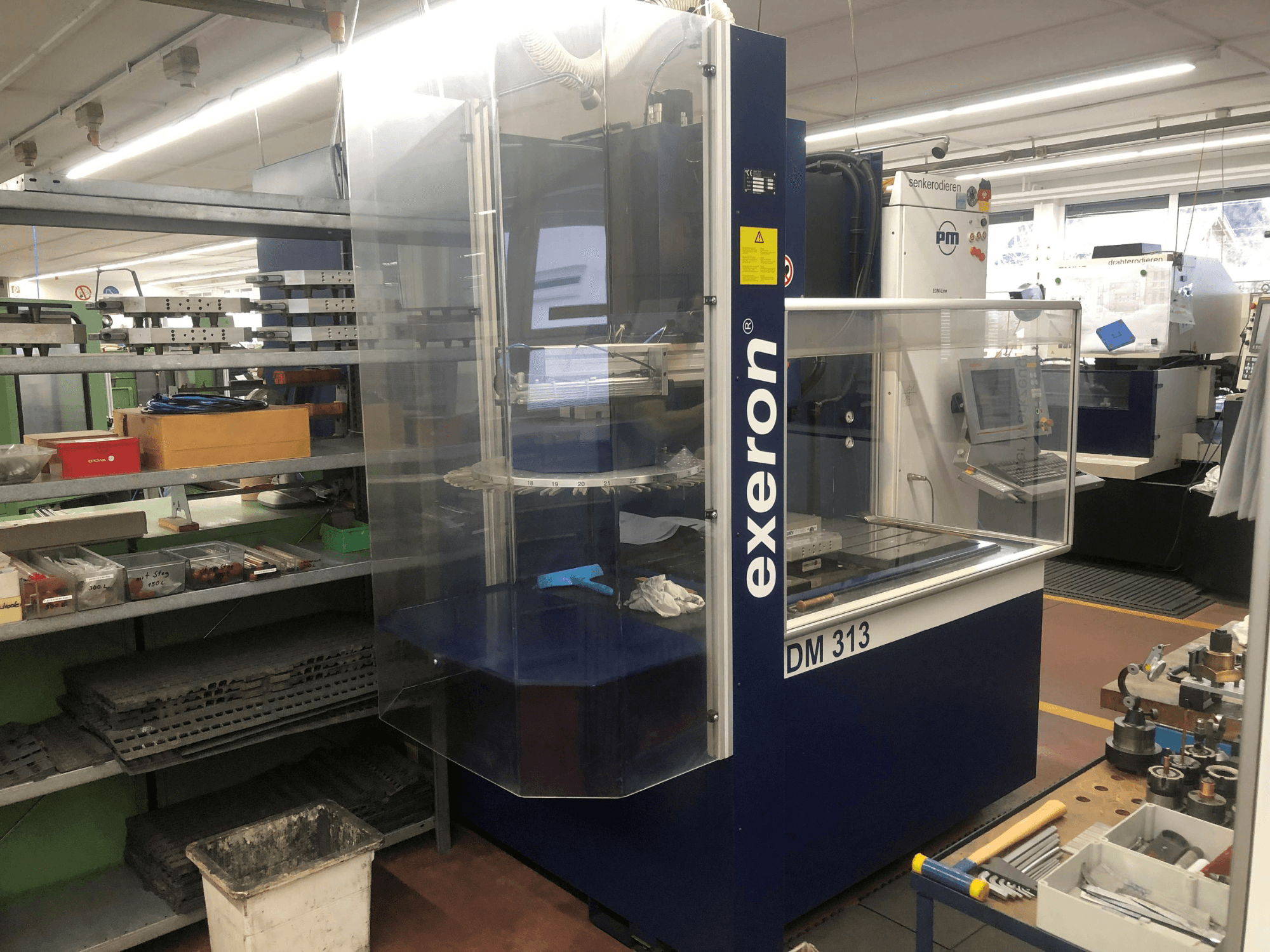 Máquina de troquelado Exeron EDM 313 en un taller, mostrando la vista frontal con carcasa transparente y panel de control.