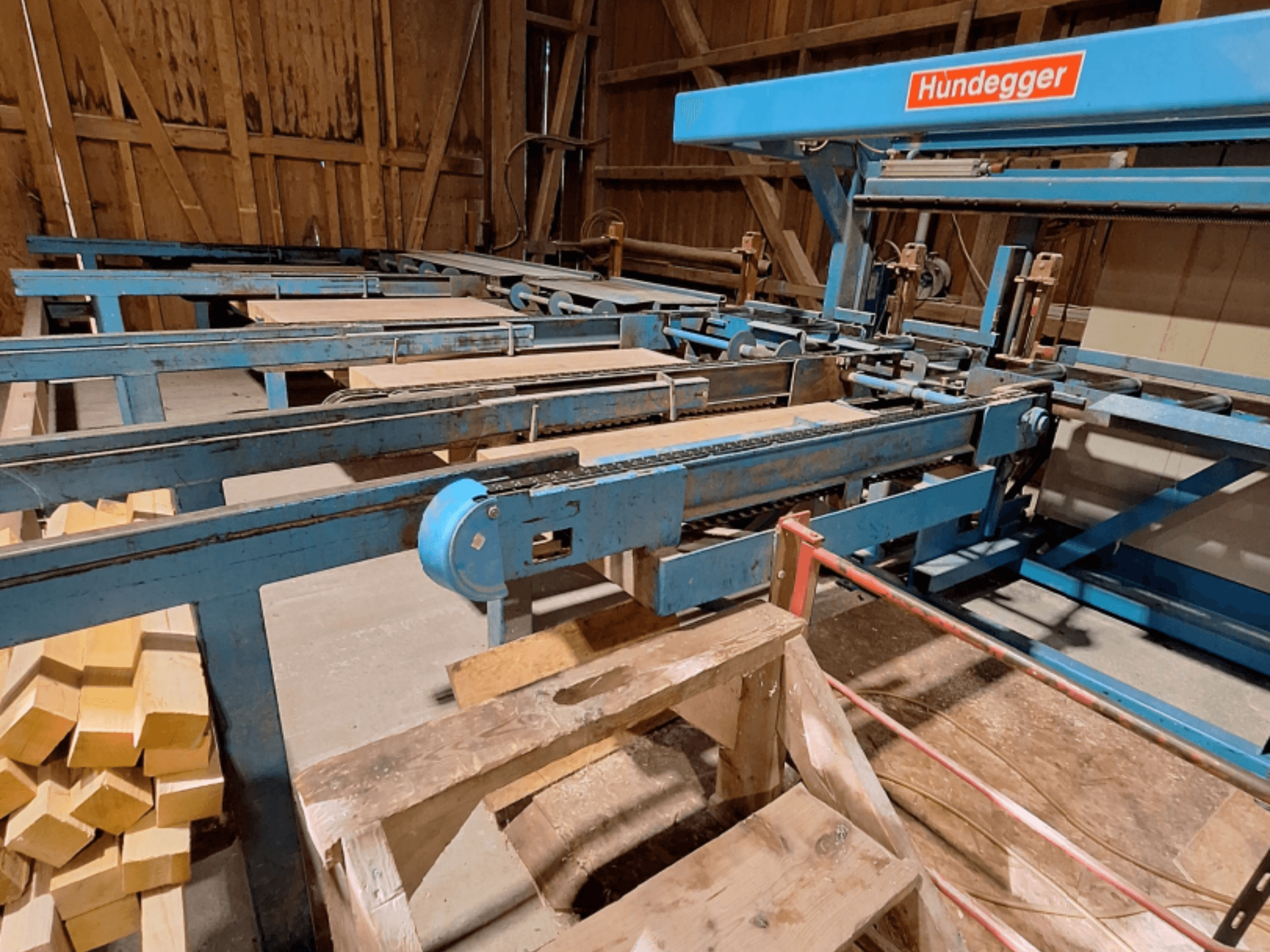 Centro de mecanizado de madera CNC HUNDEGGER K2-A en un granero, con estructura metálica azul, sistema de cinta transportadora y vigas de madera apiladas.