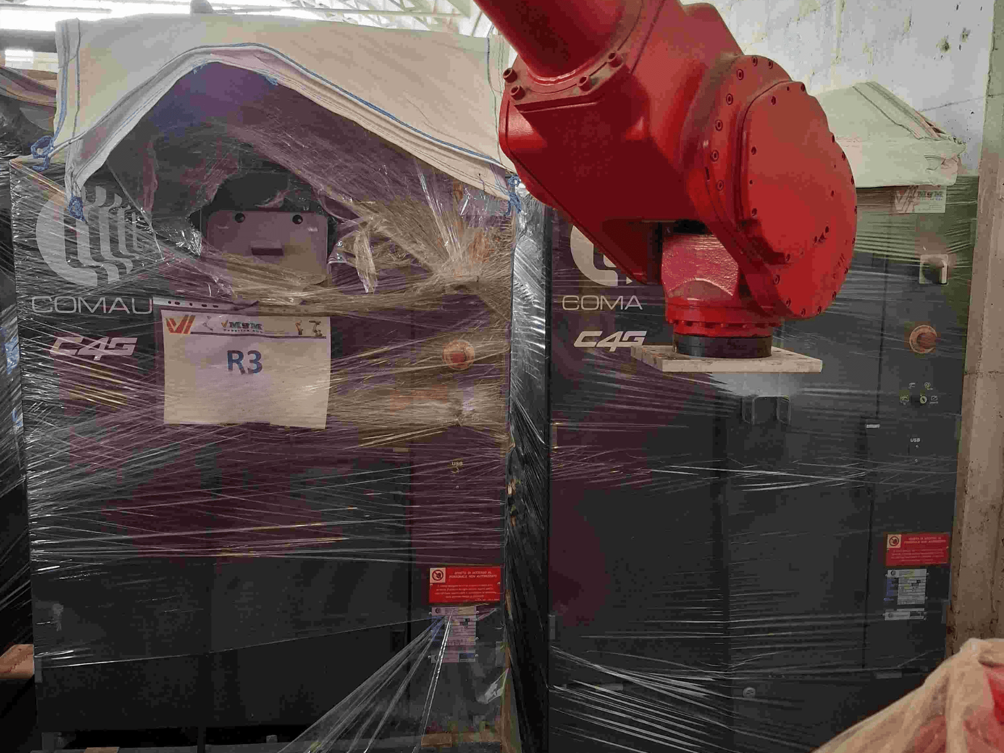 Brazo robótico Comau C4G visto de frente, envuelto en plástico, con la etiqueta "R3" y pegatinas de precaución visibles en la máquina.