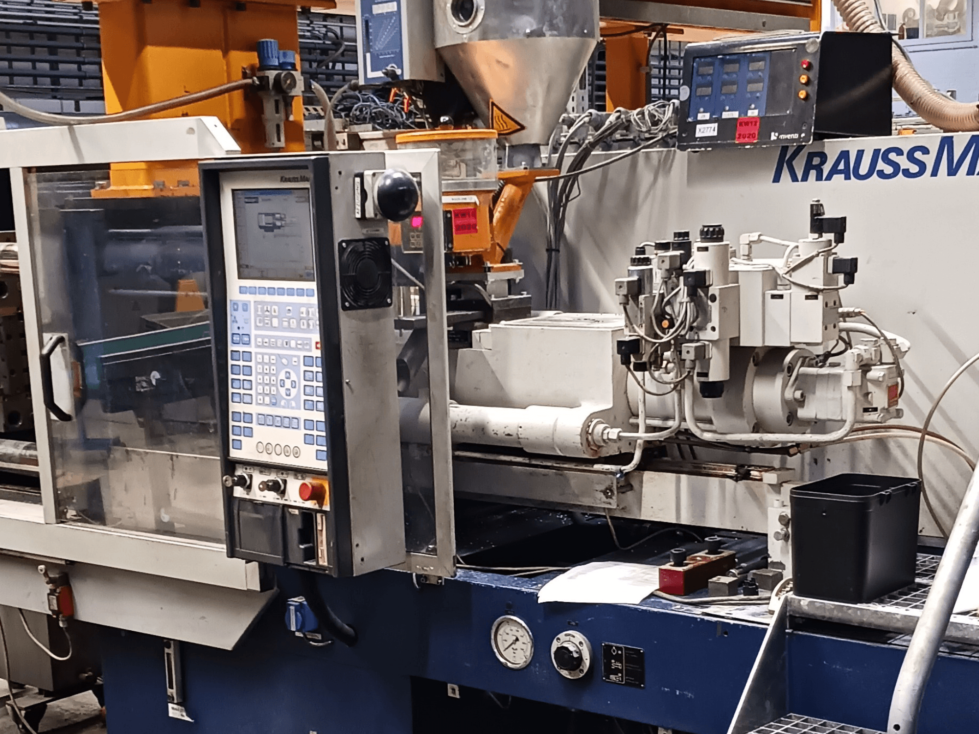 Máquina de moldeo por inyección KraussMaffei con panel de control, unidades hidráulicas y herramientas en primer plano, vista frontal.