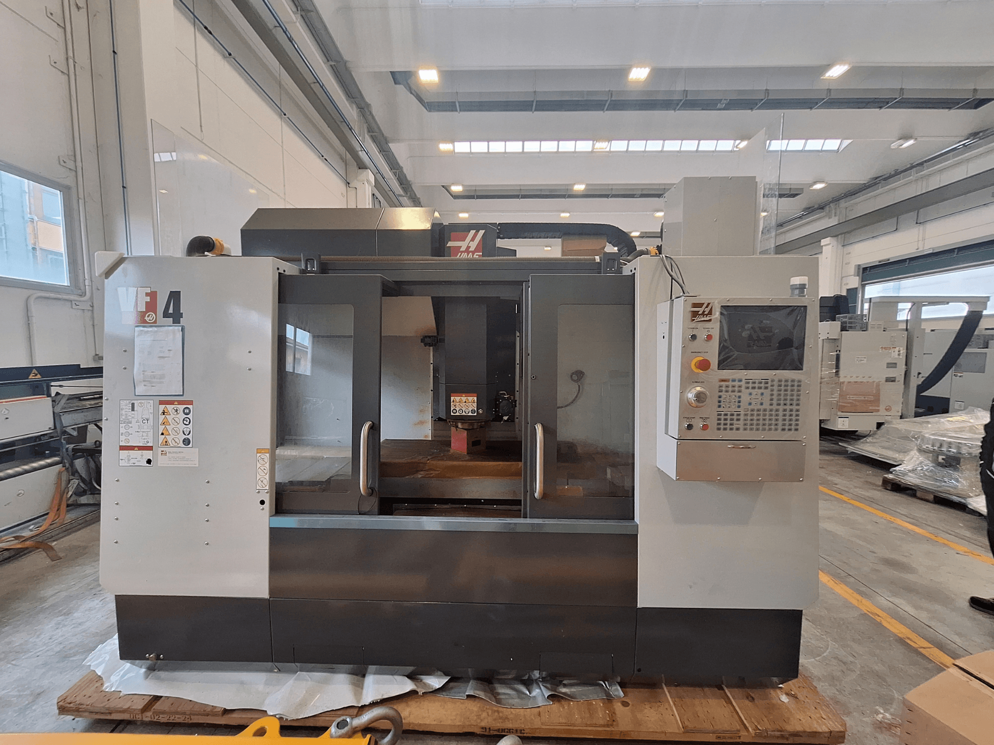 Centro de mecanizado vertical HAAS VF-4, vista frontal, en la que se aprecian el panel de control, la zona de trabajo y las etiquetas de seguridad en un entorno de almacén.