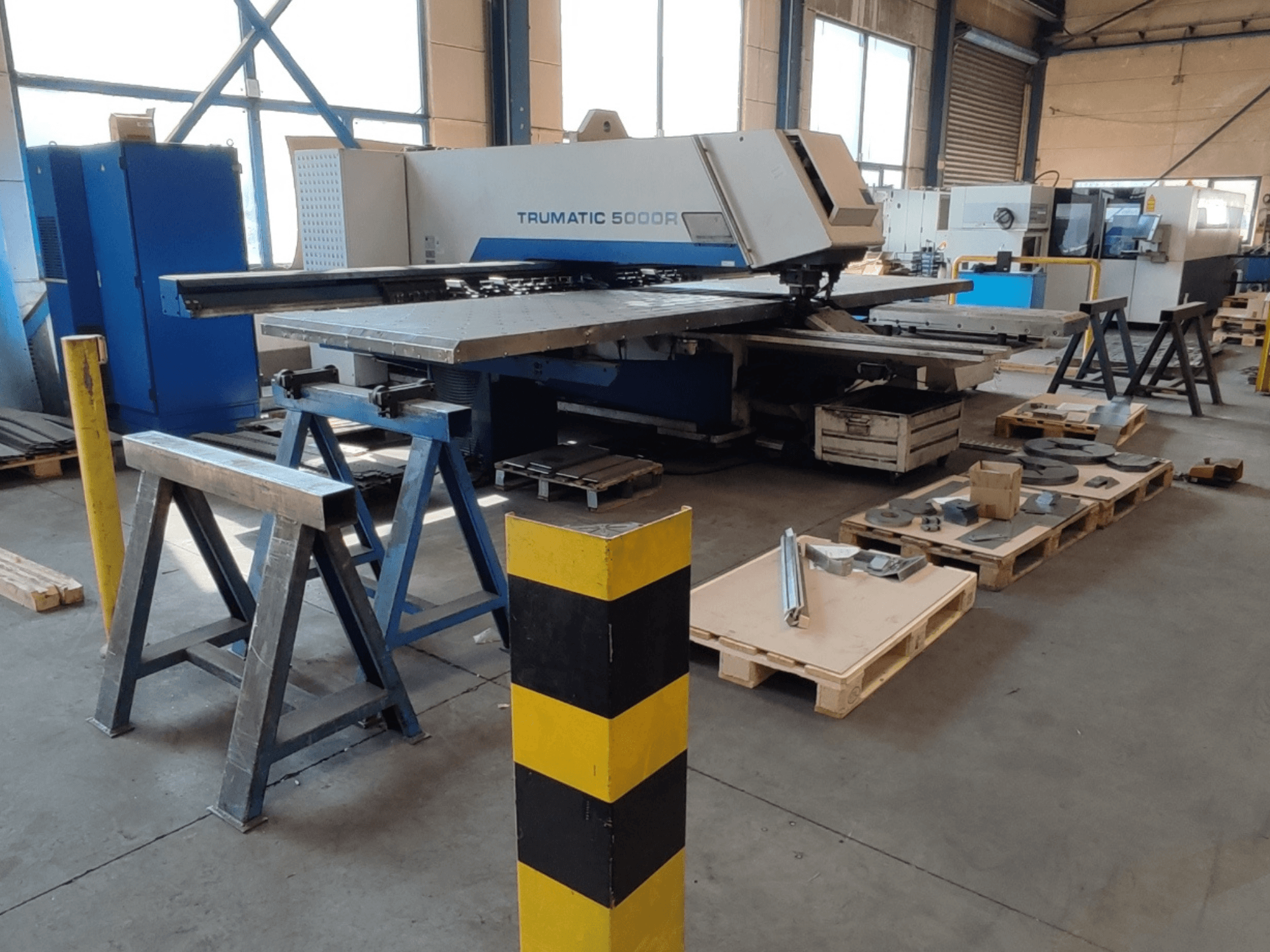 Punzonadora CNC TRUMPF Trumatic 5000 R en un taller, vista lateral con herramientas y materiales en palés a su alrededor.