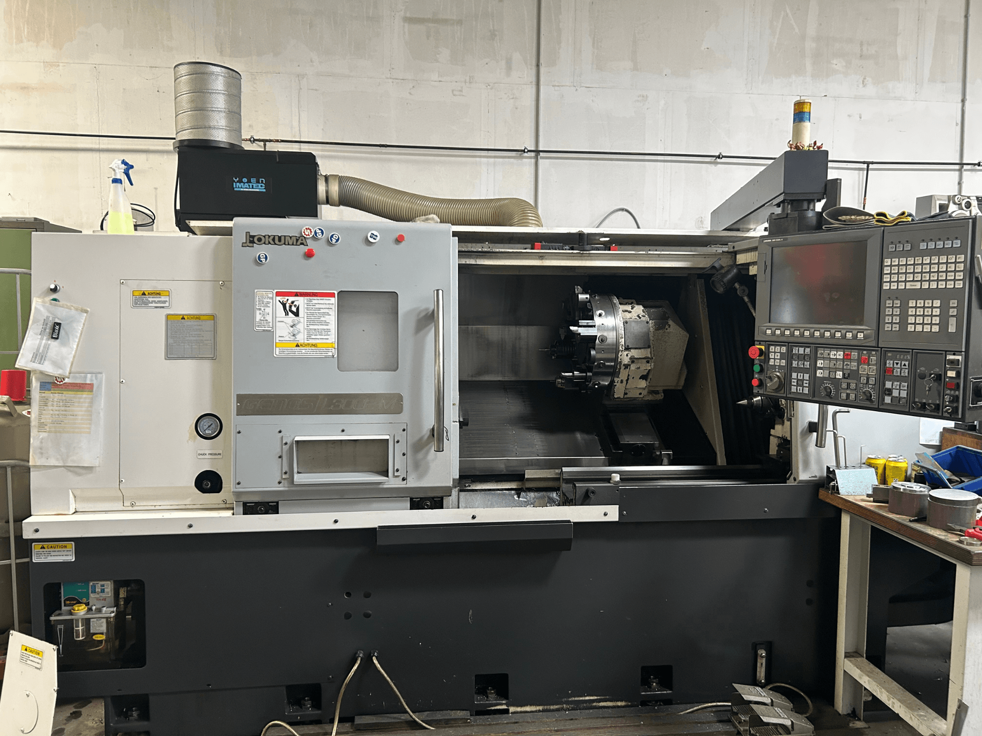 Vista frontal de un torno horizontal Okuma Genos L300E-M con panel de control, husillo visible y diversas herramientas de preparación.
