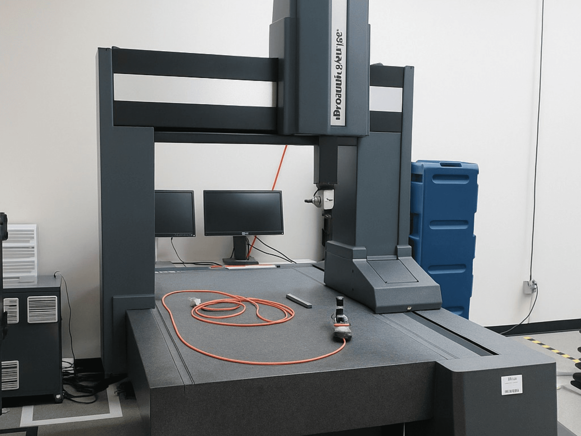 Máquina de medición de coordenadas Brooks Automation con dos monitores, vista frontal, cuenta con un tablero gris y herramientas de medición.