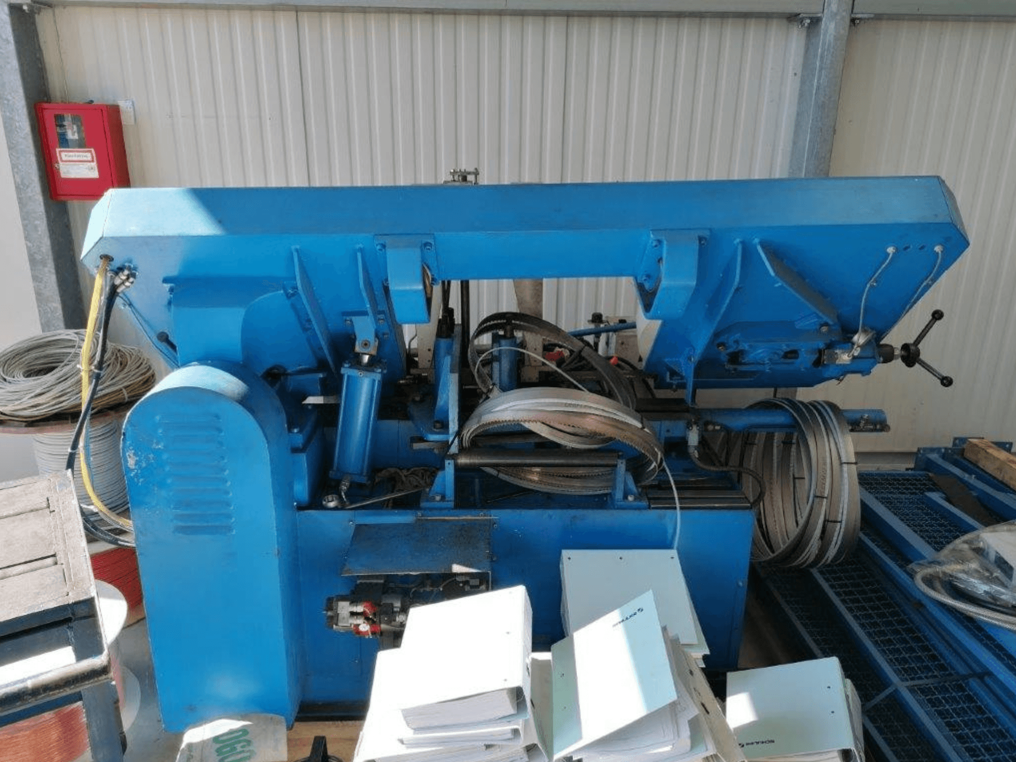 Máquina industrial azul con componentes hidráulicos, vista lateral, incluye mangueras y láminas de metal sobre una superficie.