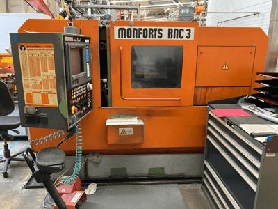 Máquina Monforts RNC 3 en naranja y negro, vista frontal, con panel de control y varios cajones para herramientas.