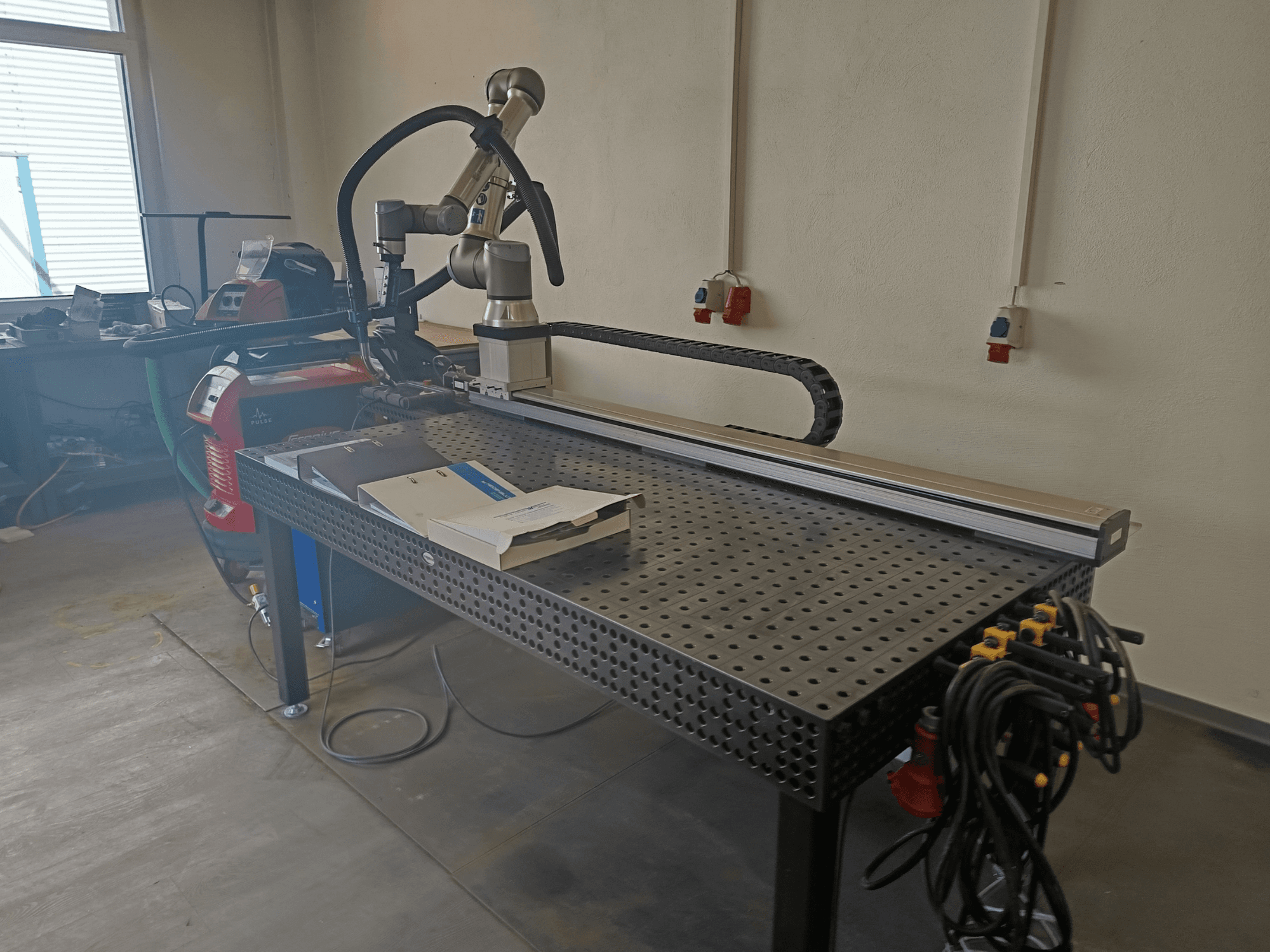 Célula de soldadura por pulsos FRONIUS TransSteel 4000 vista de perfil, con un brazo robótico, un espacio de trabajo y conexiones eléctricas.