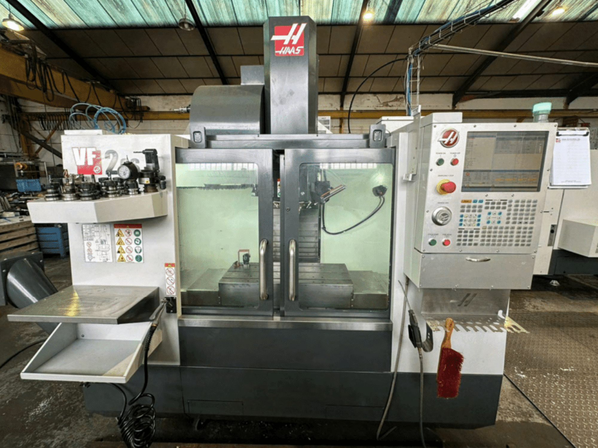 Vista frontal de la máquina CNC VF2 de Haas, mostrando el panel de control, los portaherramientas y el espacio de trabajo con etiquetas de seguridad y un cepillo.