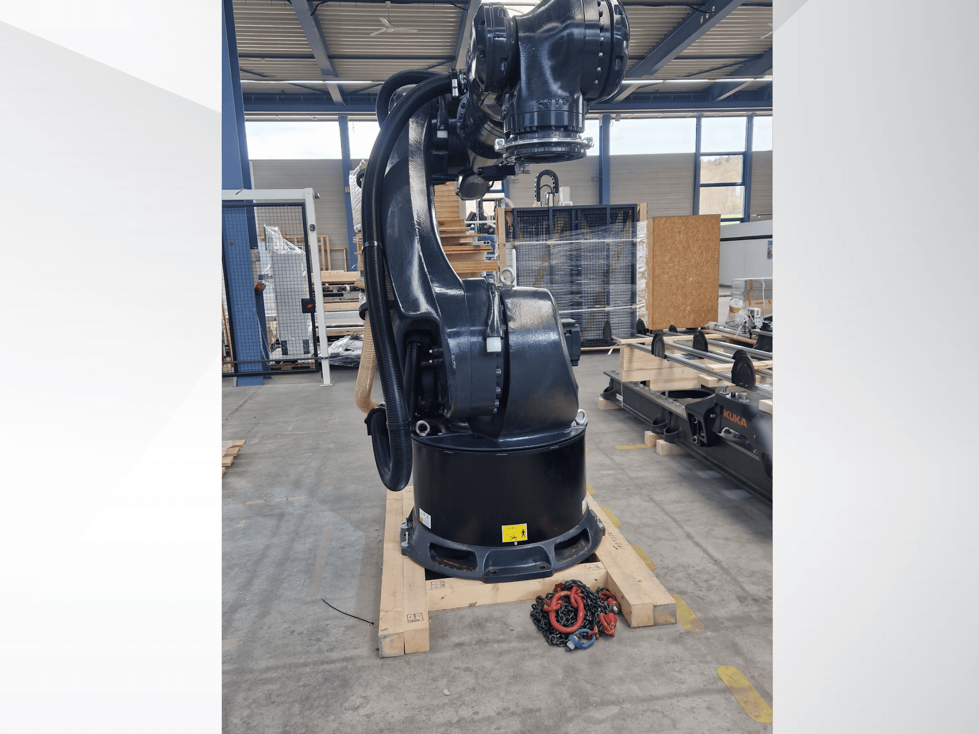 Brazo robótico KUKA KR 340 R3330 en un almacén; vista lateral en la que se aprecian su base y los cables, con cadenas en primer plano.
