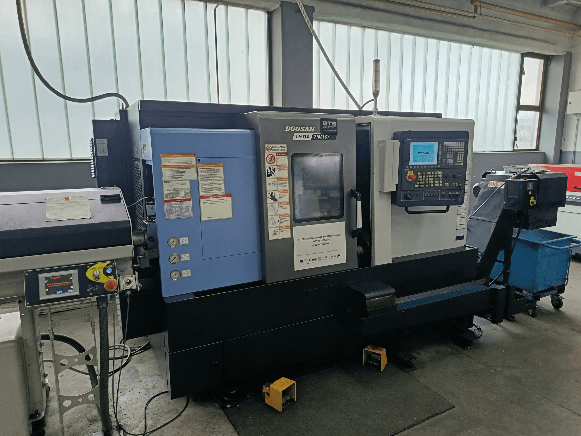 Centro de torneado y fresado DOOSAN LYNX 2100 LSY, vista lateral mostrando el panel de control y los botones operativos, con etiquetas informativas visibles.