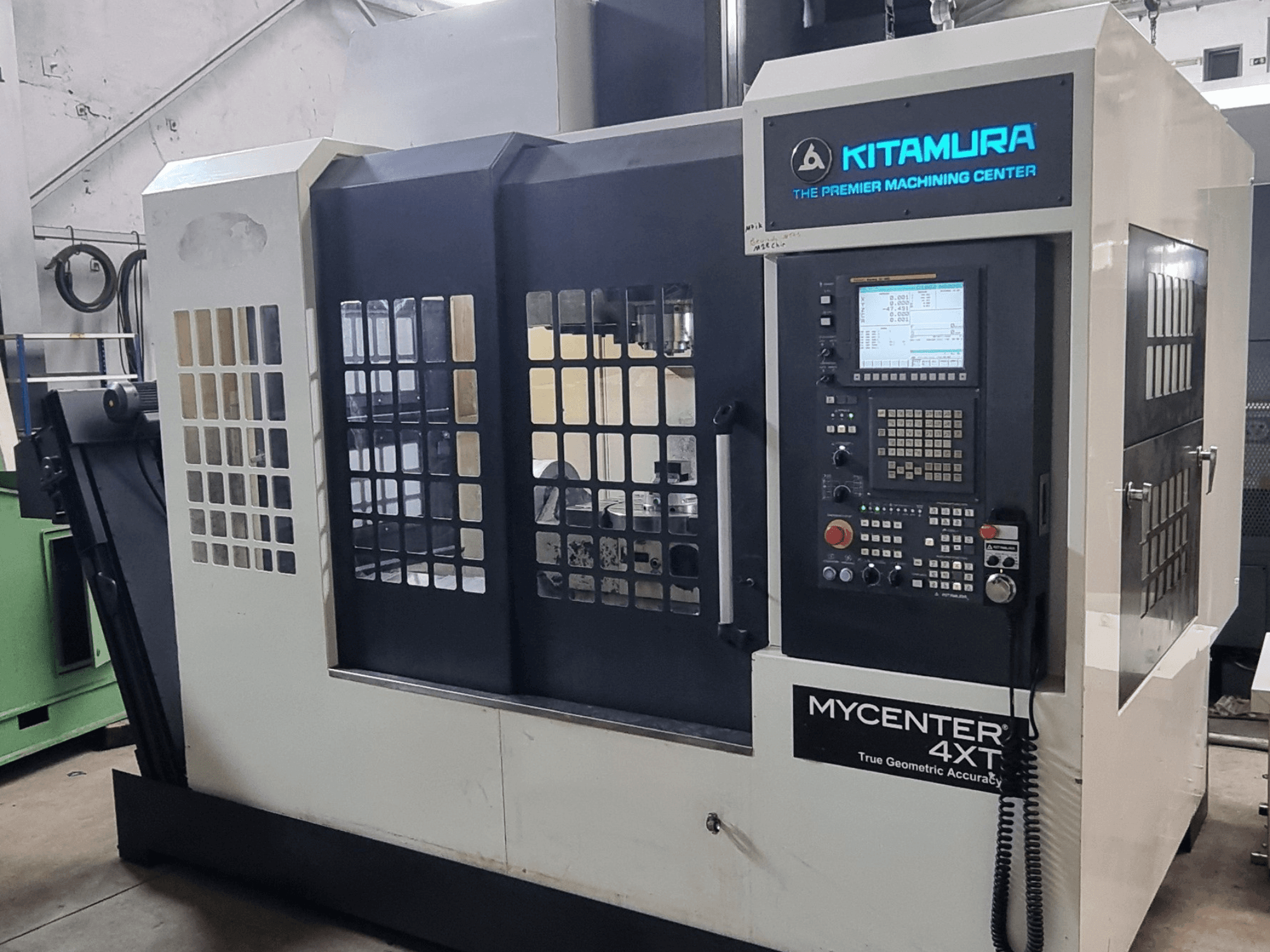 Texto alternativo: Centro de mecanizado vertical Kitamura MYCENTER 4XT, vista frontal, en la que se muestran el panel de control y los detalles de la máquina en un entorno de taller.
