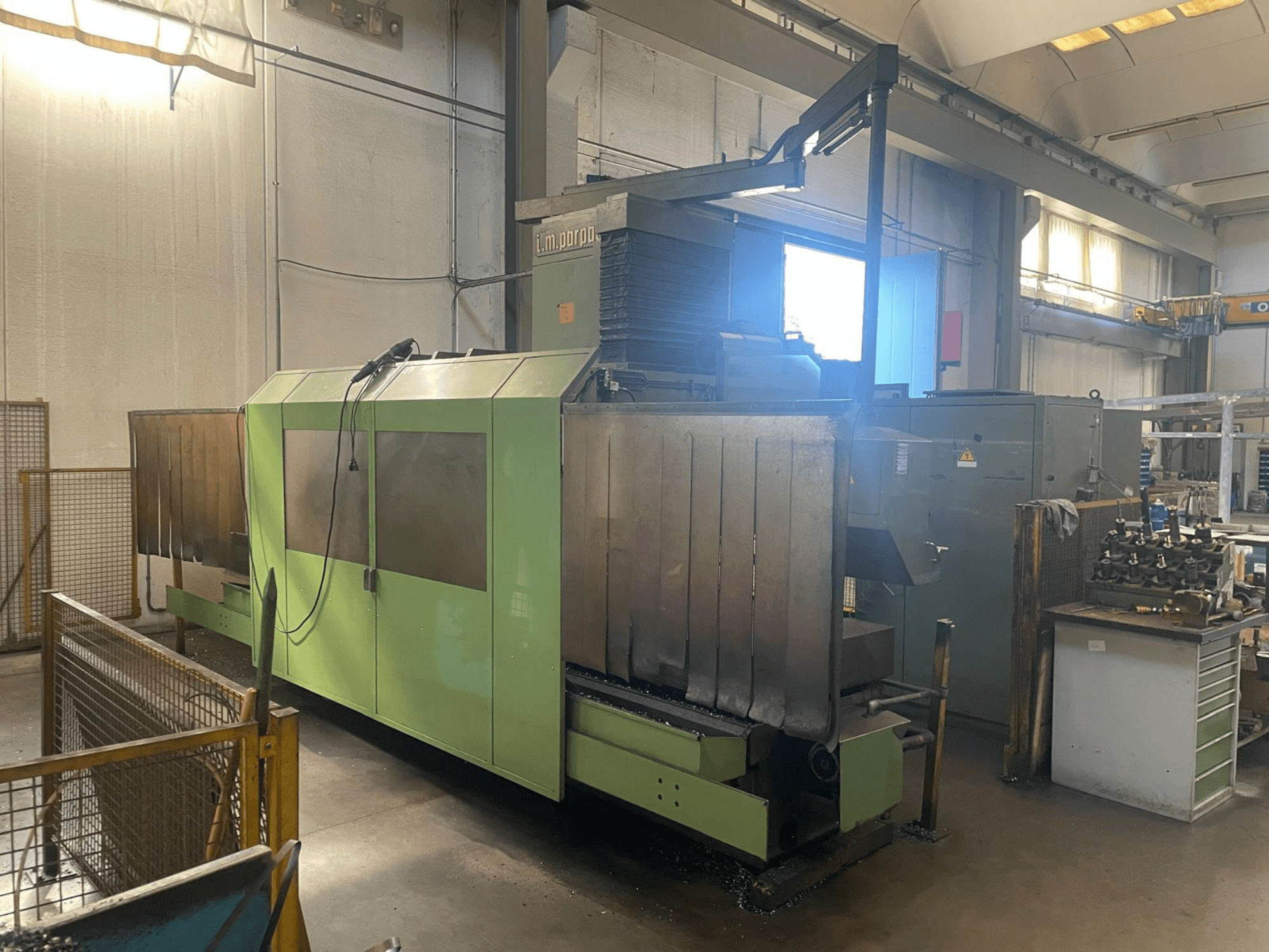 Máquina industrial verde con componentes metálicos visibles, vista frontal; cuenta con panel de control, advertencias de seguridad y estación de trabajo cercana.