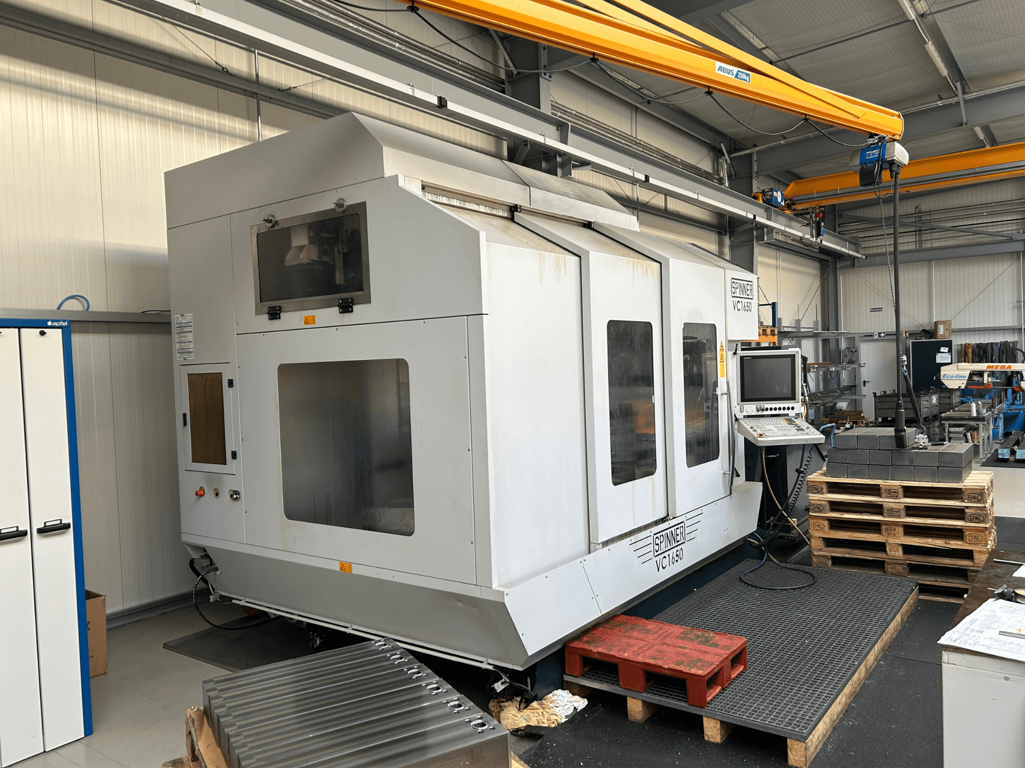 Máquina CNC Spinner VC 1650, vista frontal, con panel de control, grandes ventanas y palés apilados cerca en un entorno de taller.