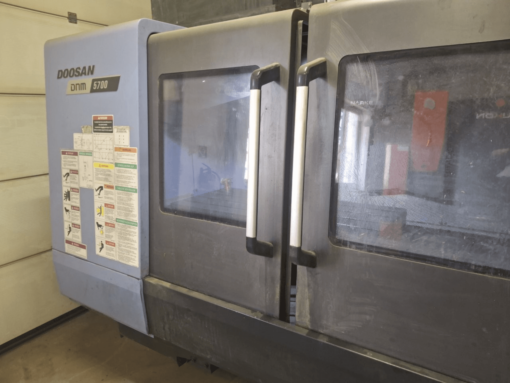 Vista frontal de un centro de mecanizado vertical DOOSAN DNM 5700, que muestra puertas dobles y paneles de control con directrices operativas.