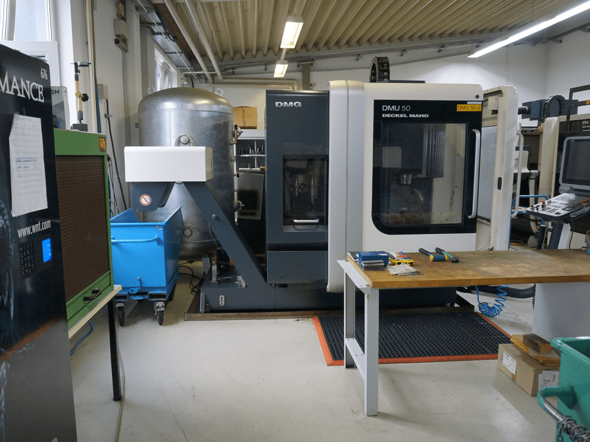 Fresadora DMG DMU 50 en un taller, vista frontal, con herramientas sobre una mesa de madera y un contenedor azul con ruedas cerca.