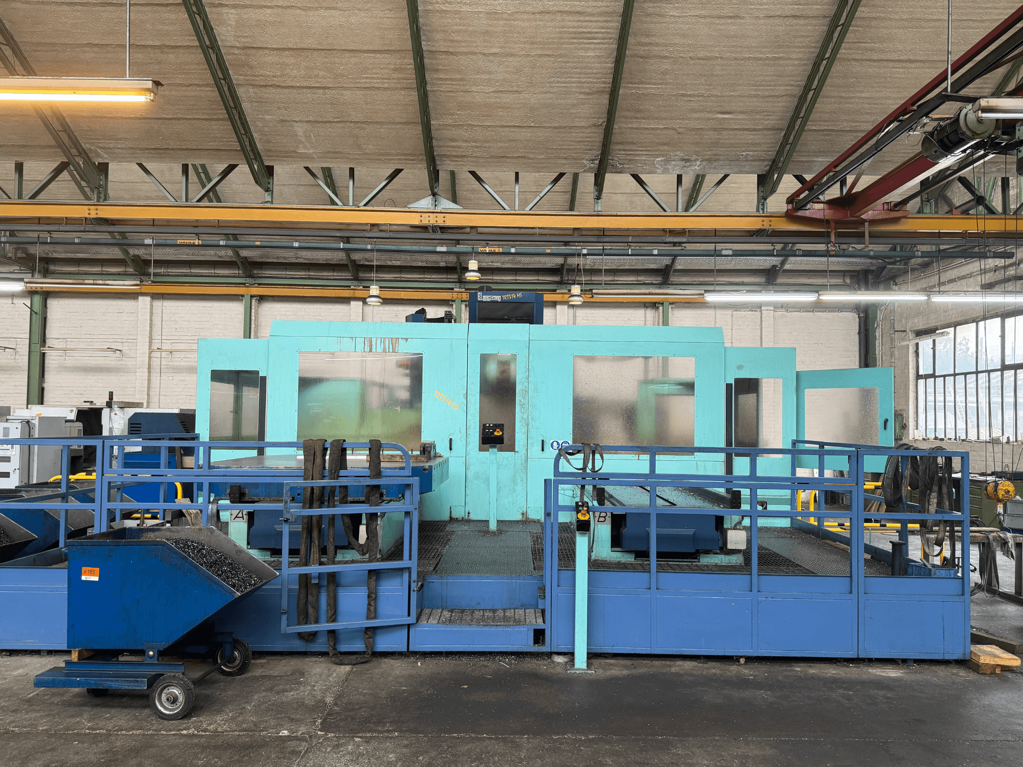 Máquina CNC industrial azul con paneles protectores, vista frontal, con almacenamiento de herramientas y un carro de recogida de residuos en el lateral.