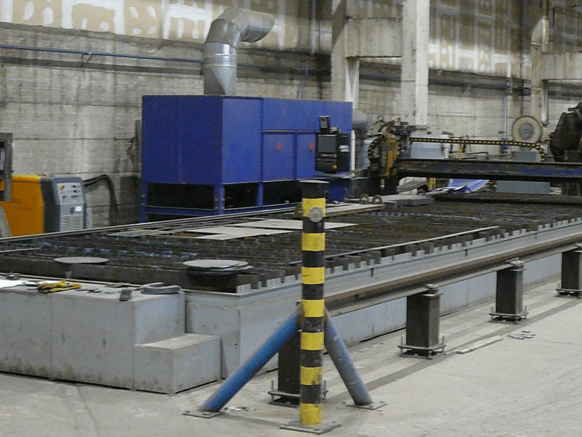 Configuración de máquina industrial de corte de metal, vista lateral, con caja de control azul, bandas de seguridad y superficie de trabajo metálica.