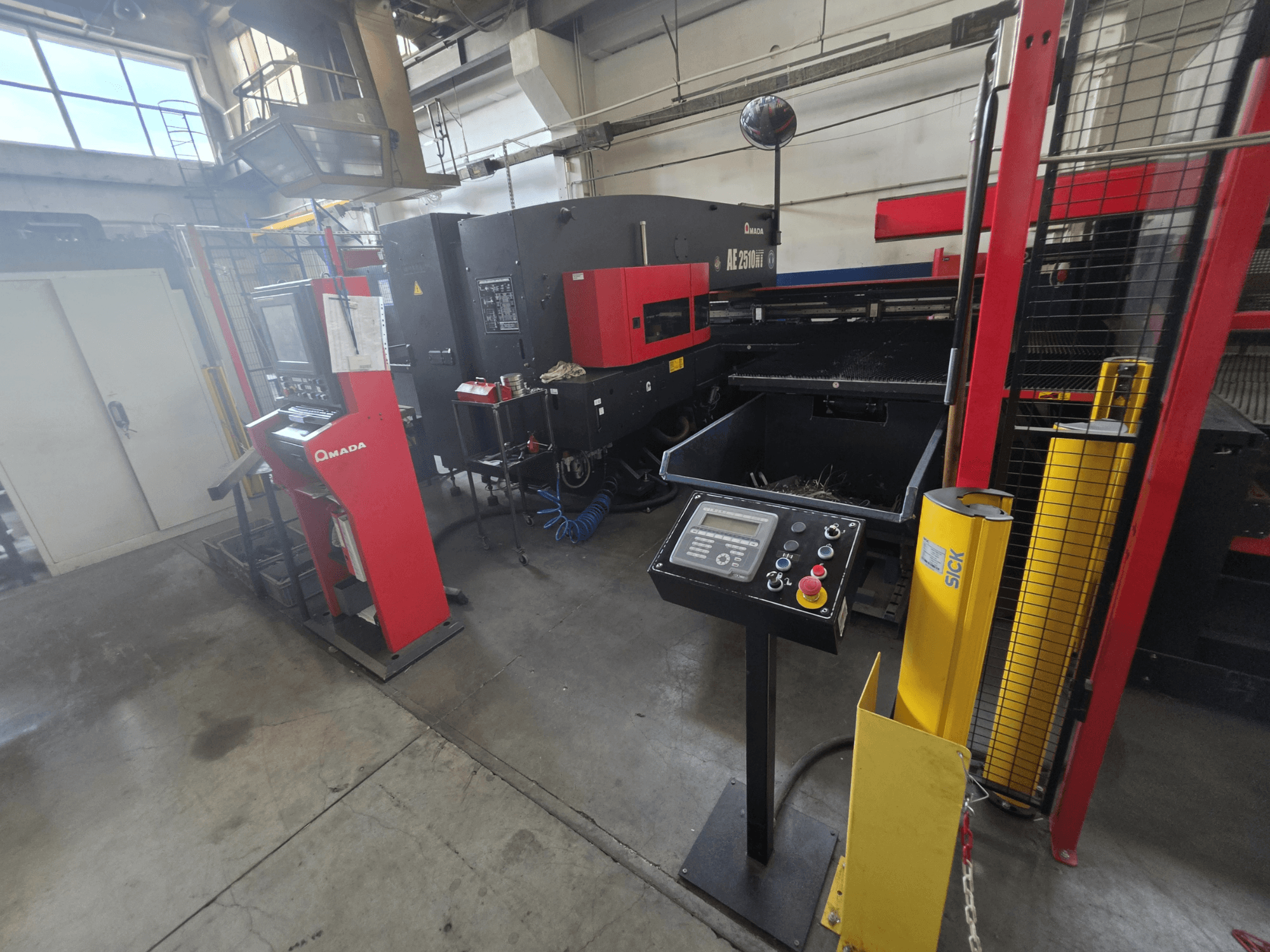 Punzonadora CNC AMADA AE-2510NT, vista frontal con el panel de control, los dispositivos de seguridad y el entorno industrial que la rodea.