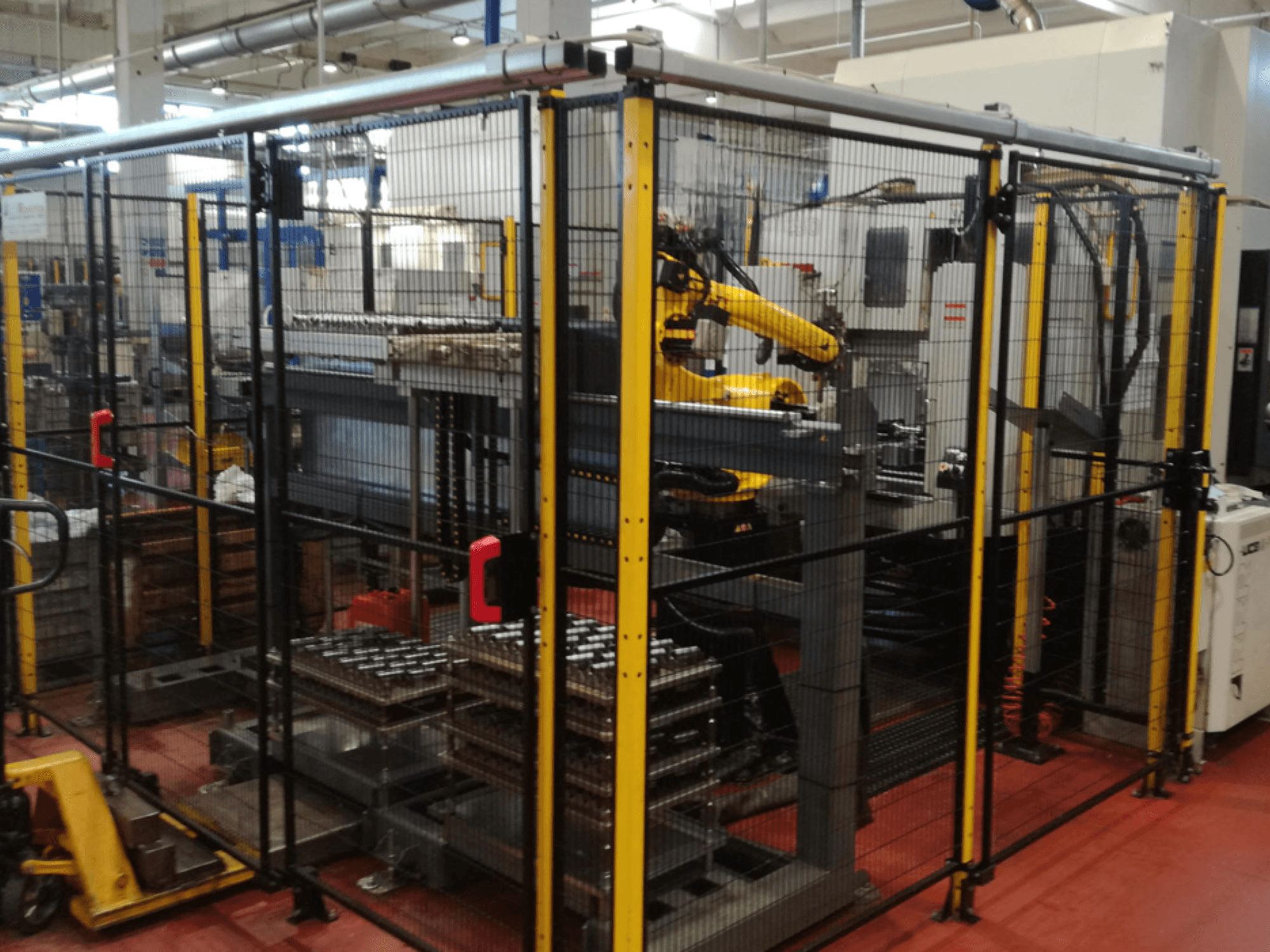Centro de mecanizado vertical Enshu EV450 en un espacio de trabajo vallado, con brazos robóticos y múltiples piezas de trabajo apiladas. Vista frontal.