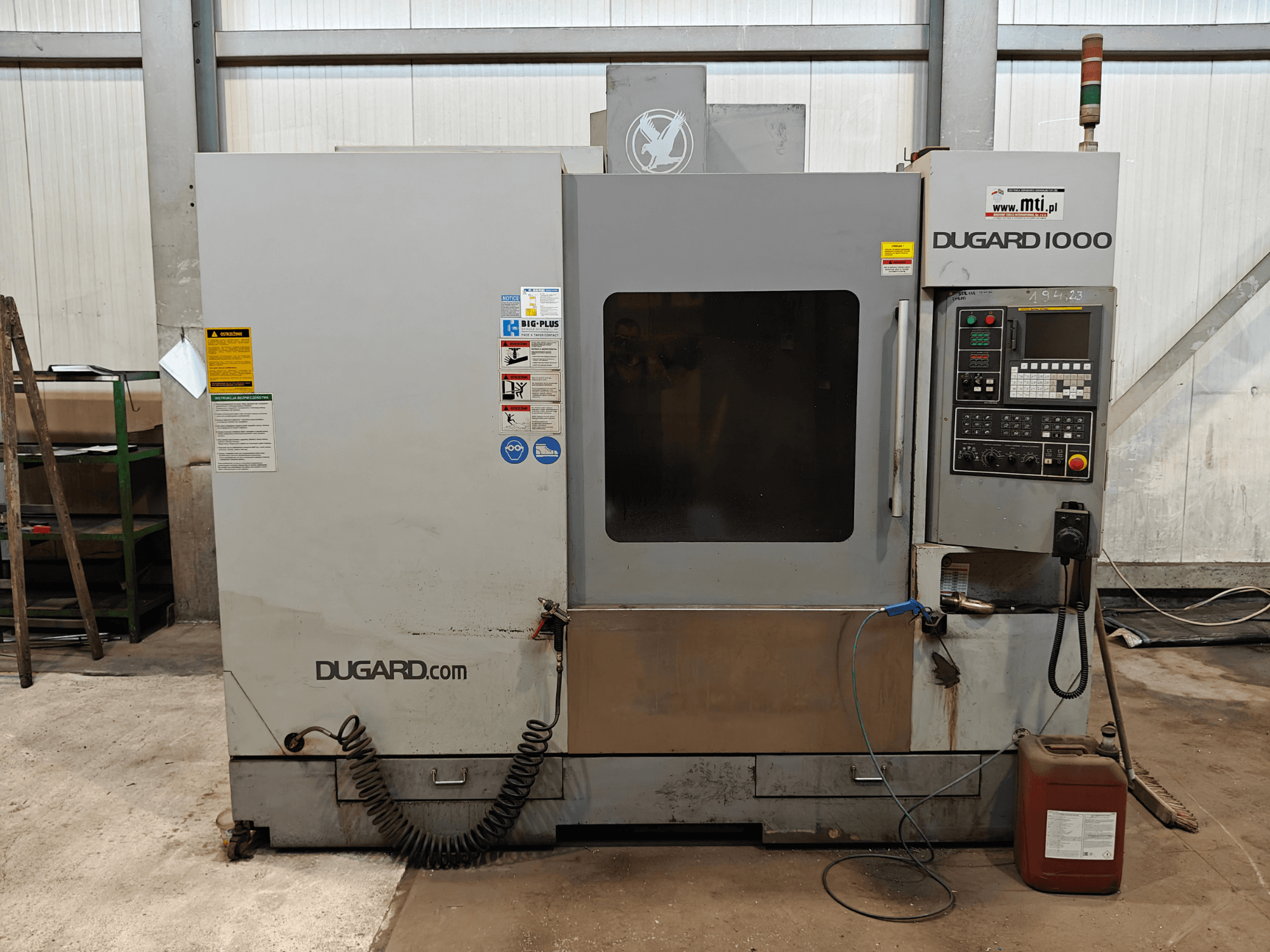 Máquina CNC Dugard 1000, vista frontal, mostrando el panel de control, las etiquetas de seguridad y el recipiente de aceite, en un entorno de taller.