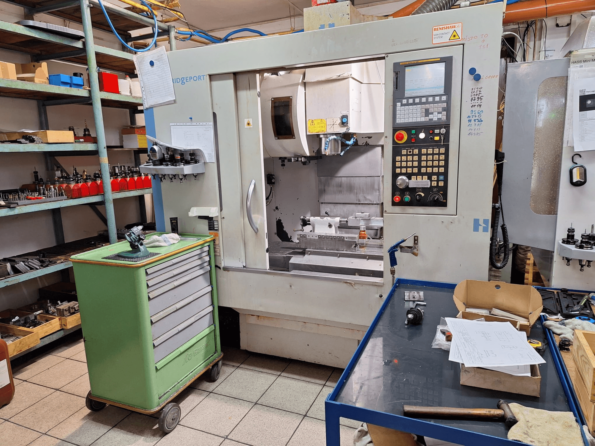 Vista frontal del centro de mecanizado vertical Hardinge Bridgeport GX 710 con panel de control, herramientas y estanterías en un entorno de taller.