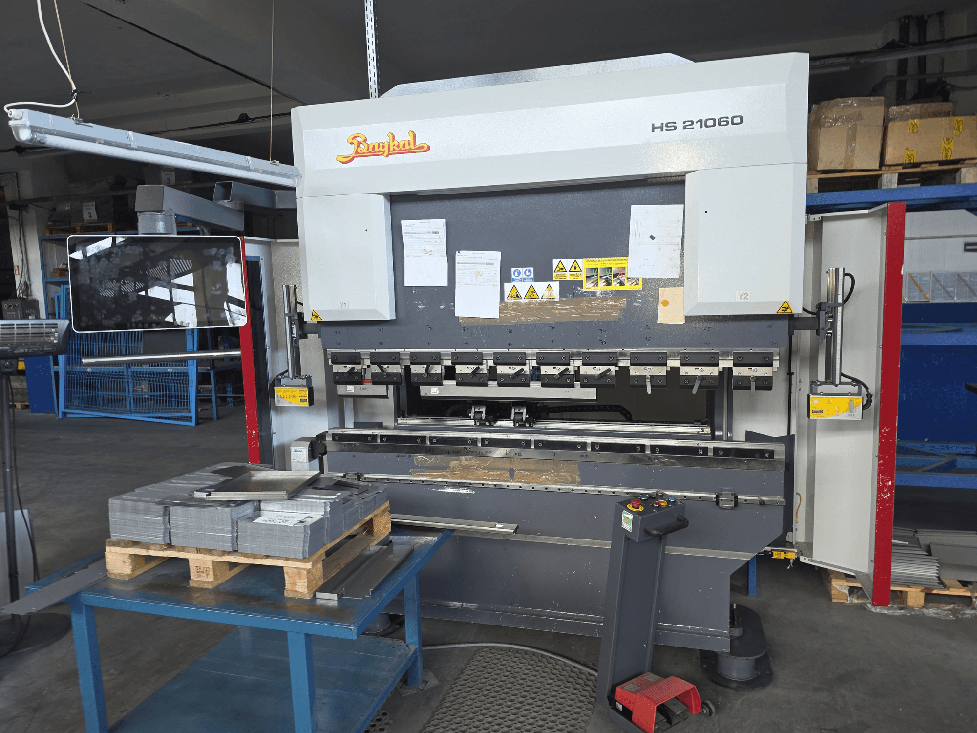 Plegadora Baykal APHS 21060 en un taller; vista lateral en la que se aprecian el panel de control, las chapas metálicas apiladas y la señalización de seguridad.