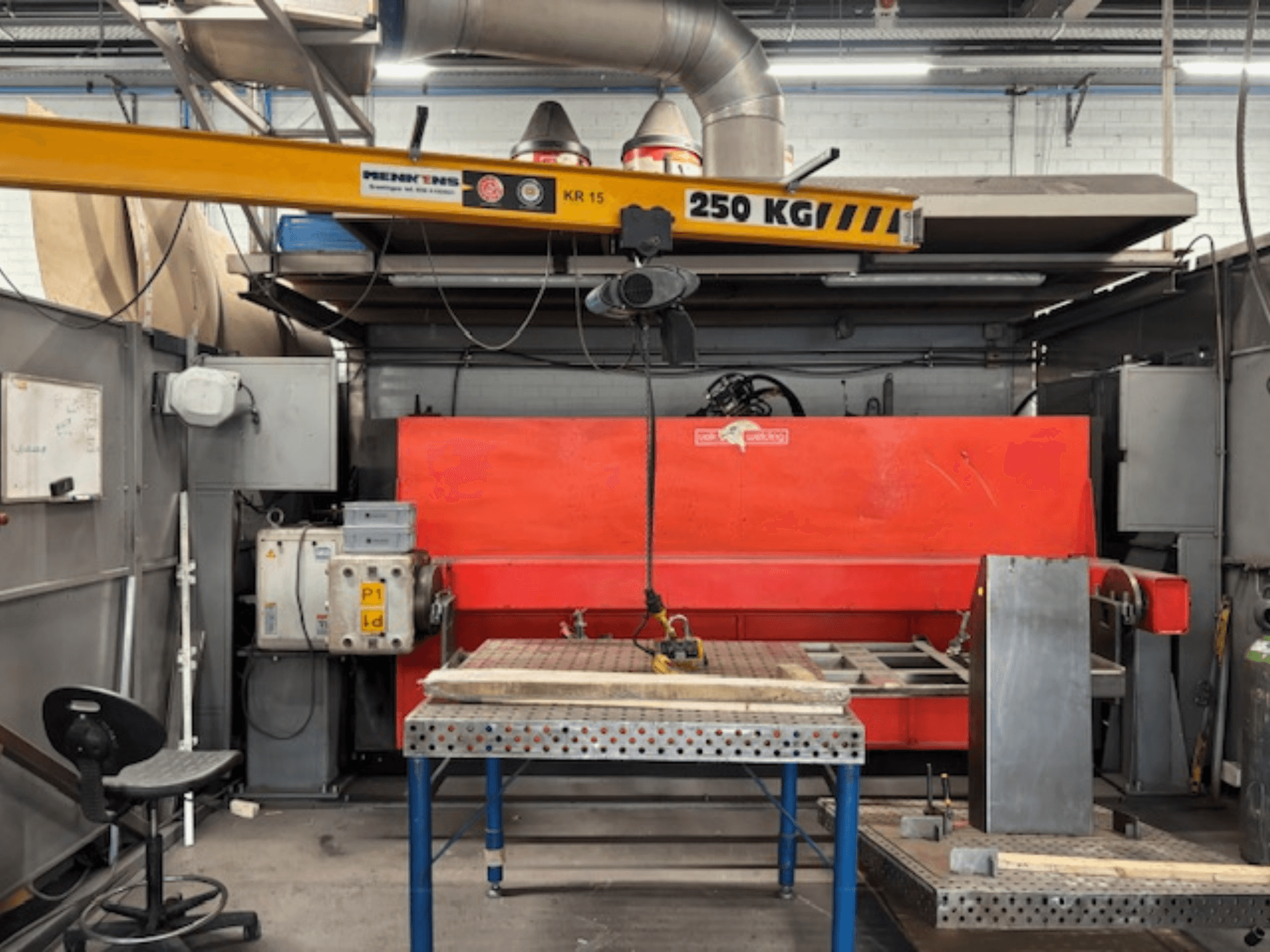 Taller industrial con una prensa hidráulica roja, una grúa puente rotulada "KR 15" y una mesa de trabajo metálica, perspectiva lateral.