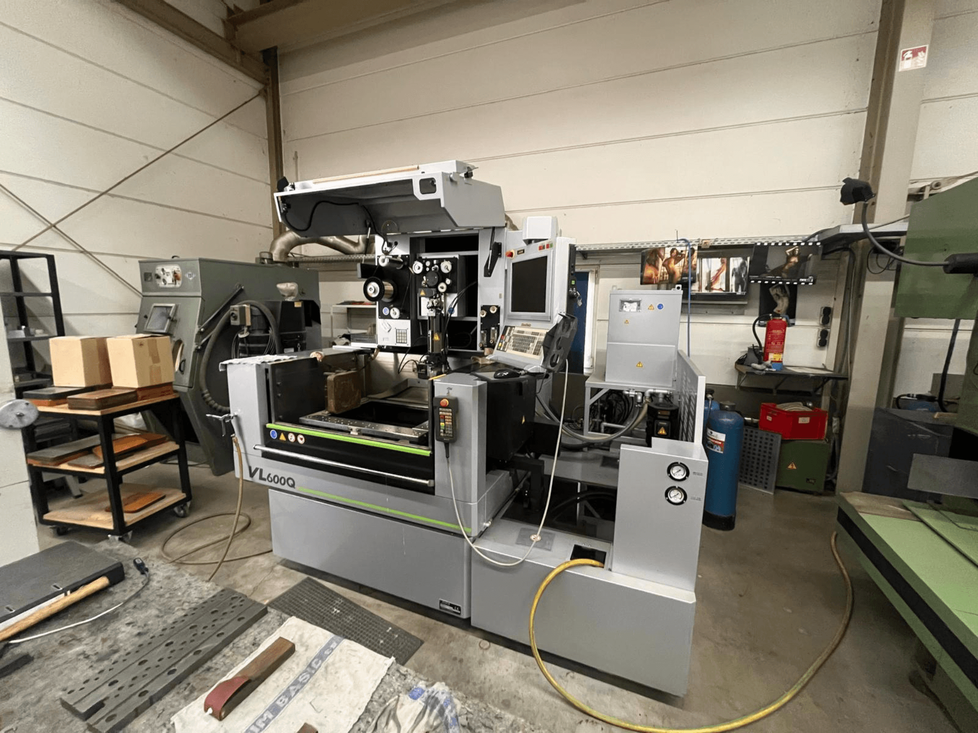 Máquina CNC VL600Q en un taller, vista frontal, con panel de control, material colocado y varias herramientas cerca.