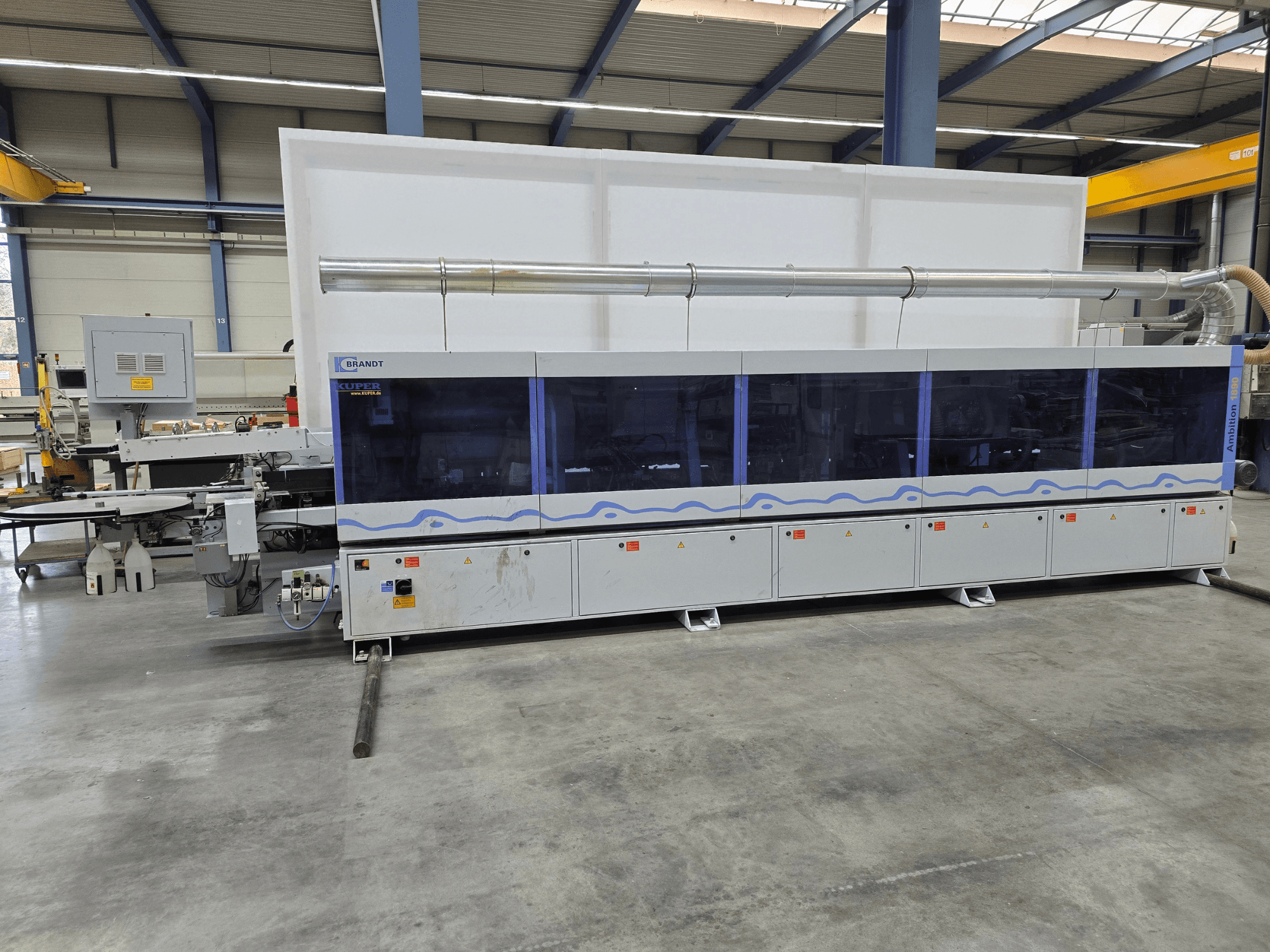 Máquina Brandt KUPER en una fábrica, vista frontal con paneles transparentes, con botones de control y advertencias de seguridad.