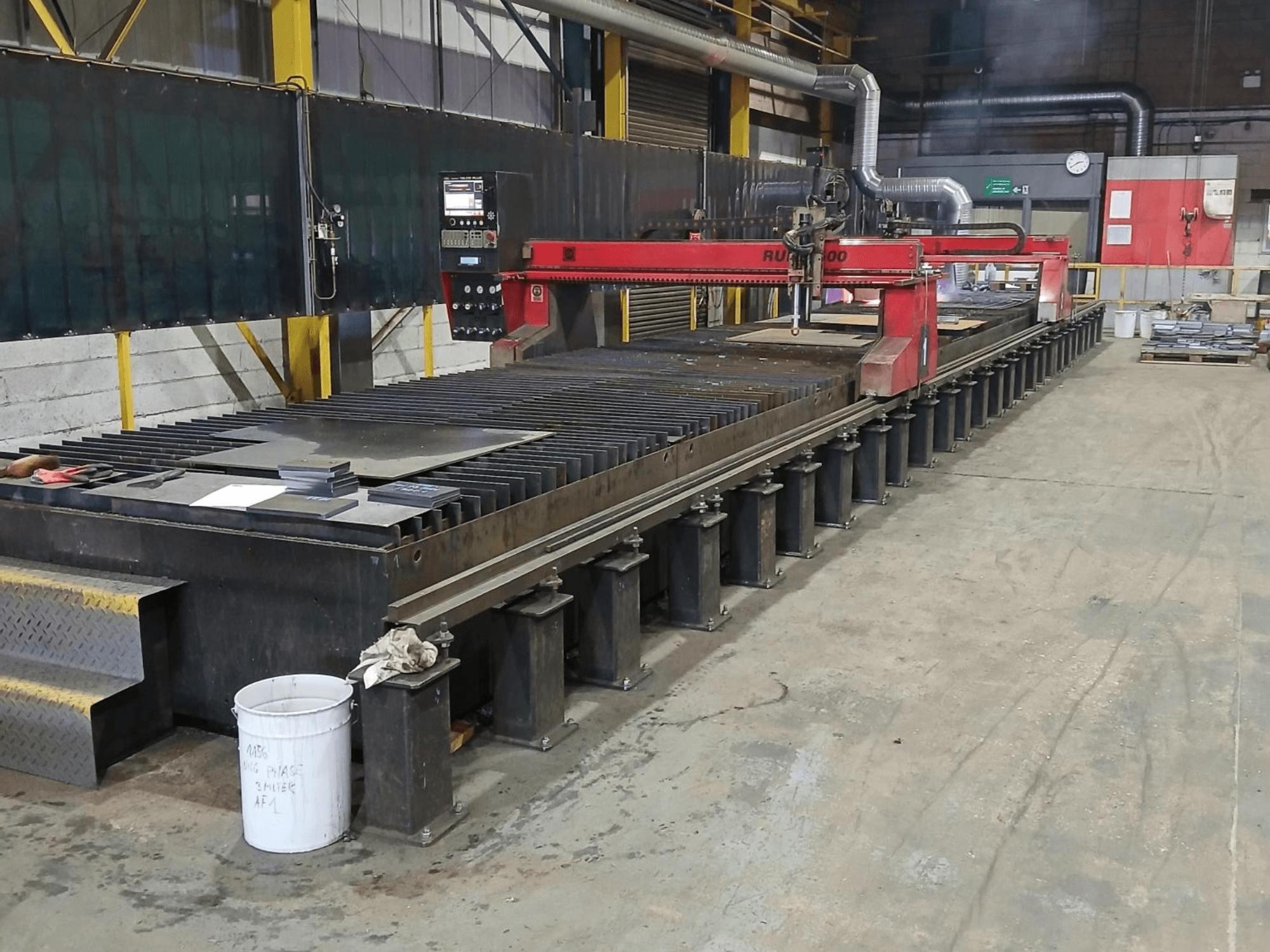 Máquina de corte RUIDA 300 roja, vista lateral, con chapa sobre una mesa de acero, situada en un espacio de trabajo industrial.