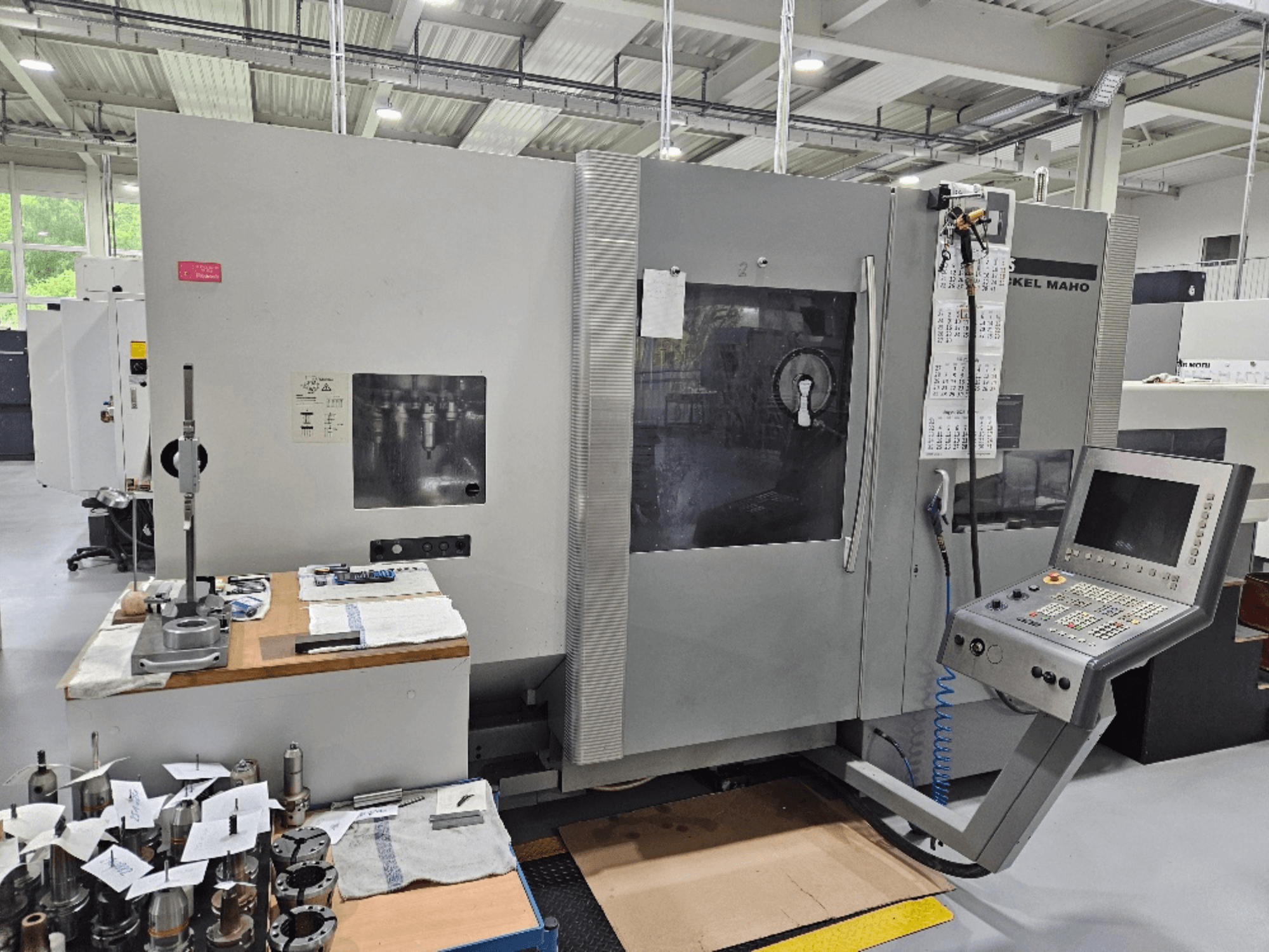 Vista frontal de una máquina DMG Mori DMU 70 con panel de control, herramientas sobre una mesa de madera y varias piezas de trabajo cerca.