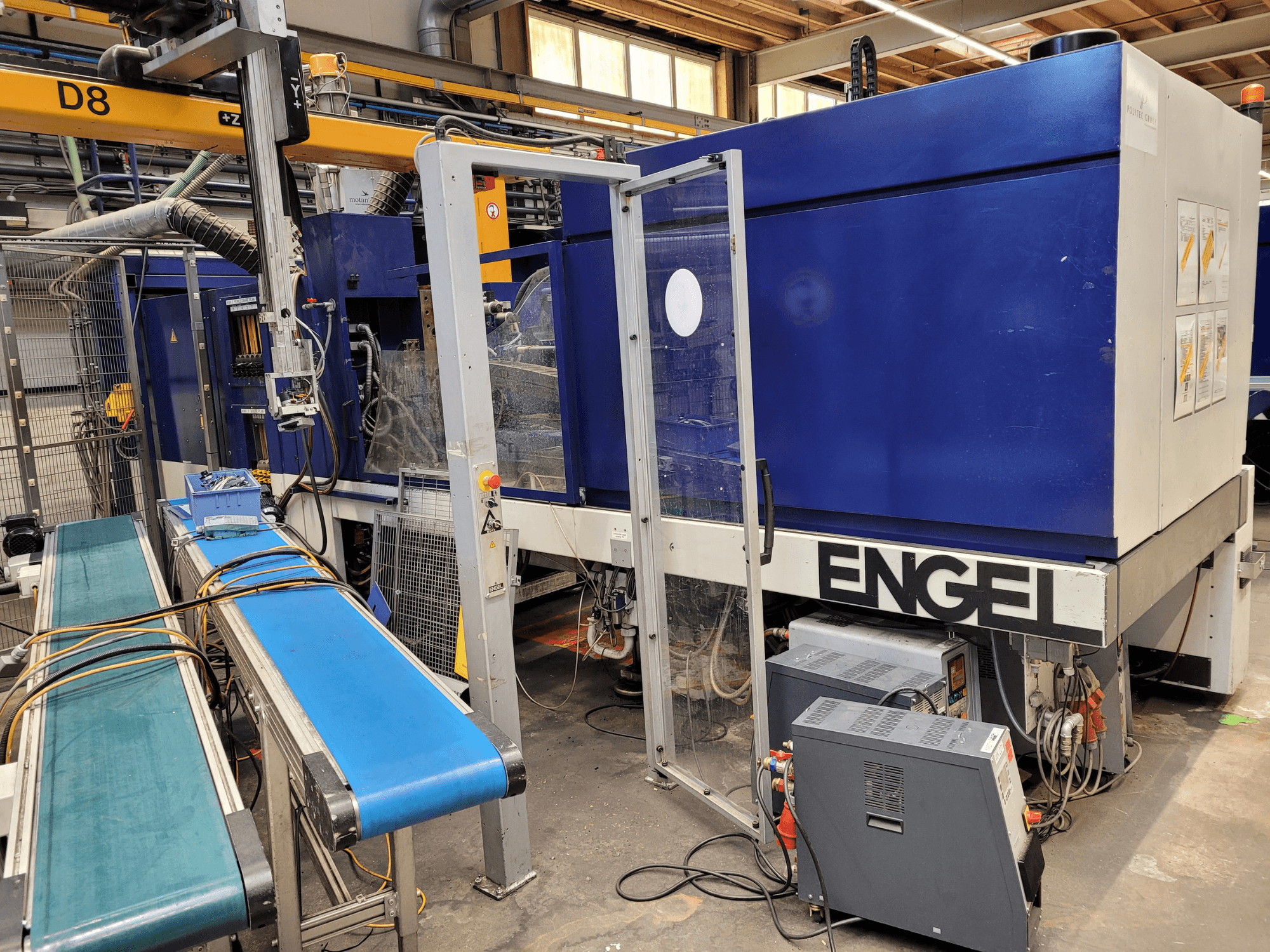 Máquina ENGEL azul con puerta de seguridad transparente, vista lateral, con panel de control, cinta transportadora y conexiones eléctricas visibles.