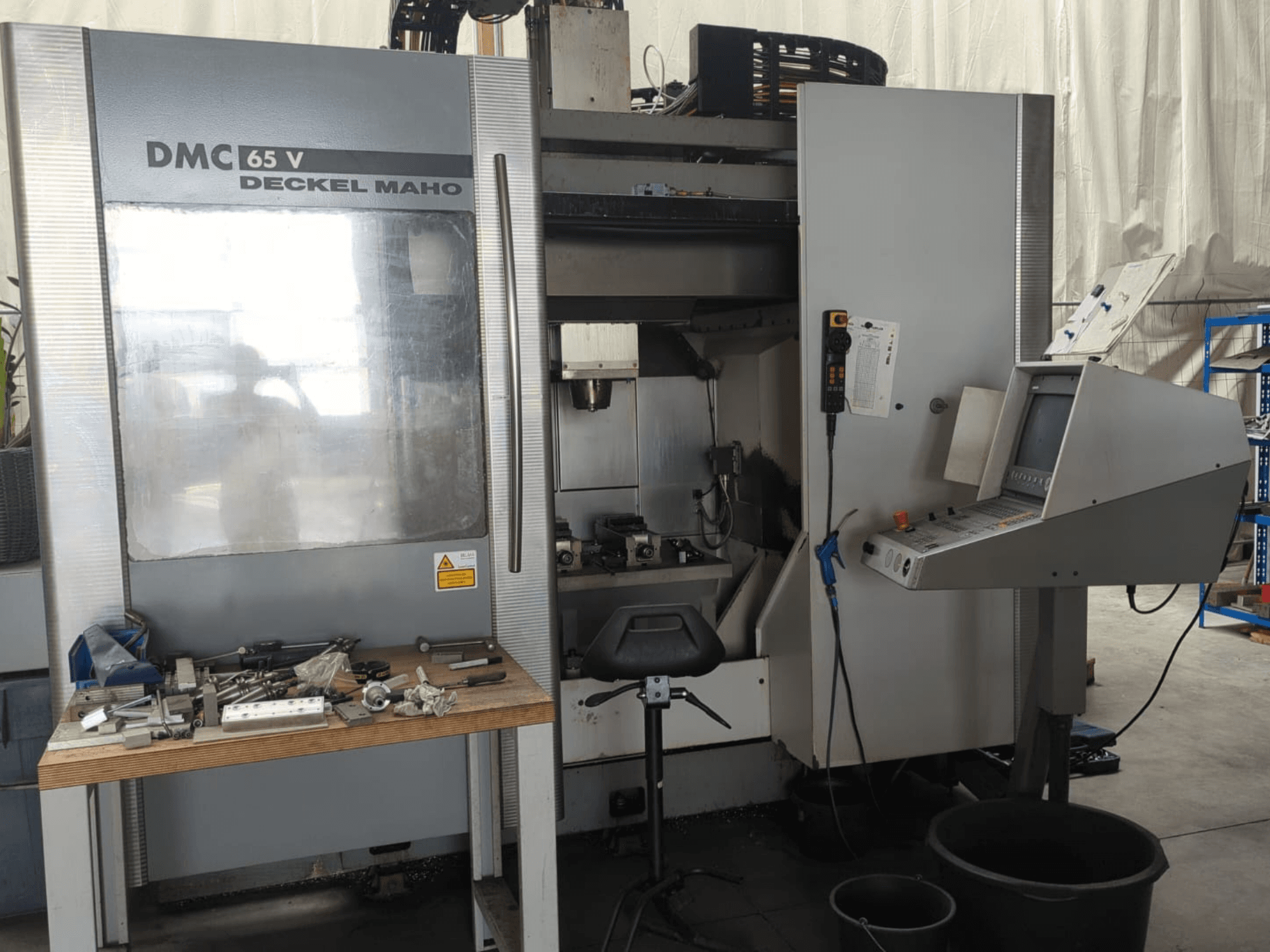 Vista frontal de un centro de mecanizado vertical DECKEL MAHO DMC 65 V con panel de control, herramientas sobre una mesa y configuración del espacio de trabajo.