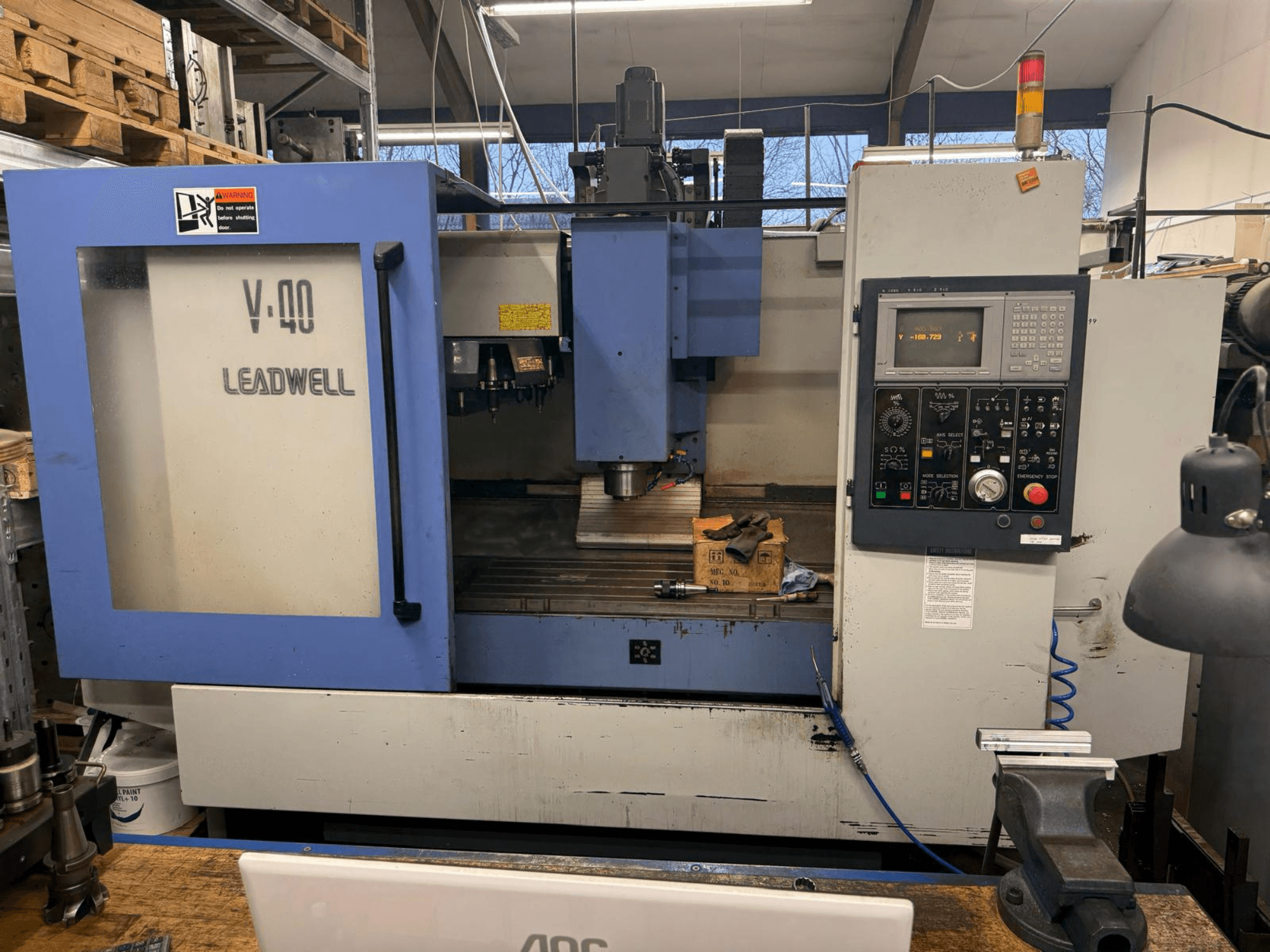 Fresadora CNC LEADWELL V-40, vista frontal, con un panel de control, herramientas de corte y un tornillo de banco sobre un banco de trabajo.