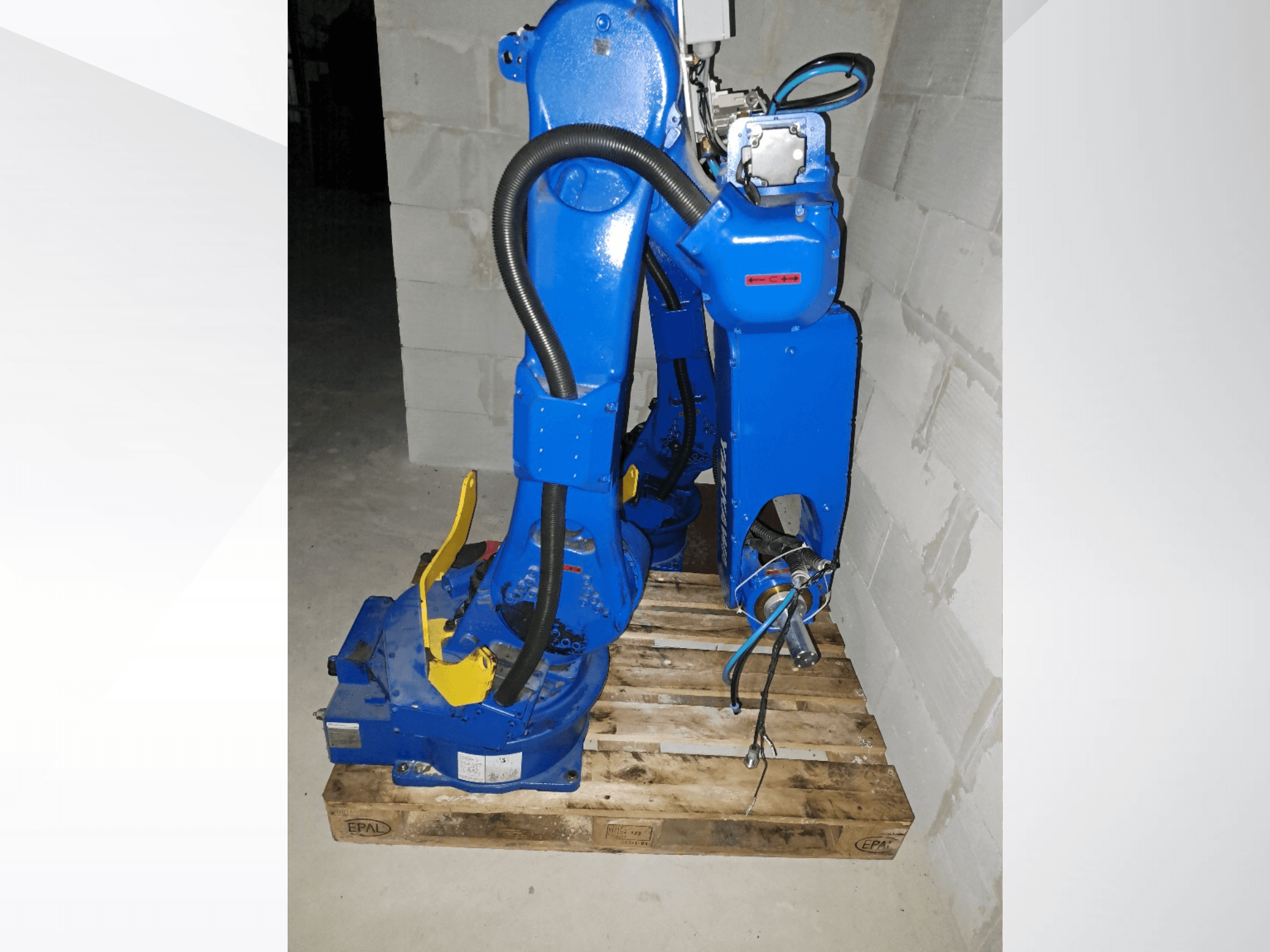 Brazo robótico Yaskawa Motoman GP25 de color azul, vista lateral sobre un palé de madera, en el que se aprecian los cables y las articulaciones mecánicas.