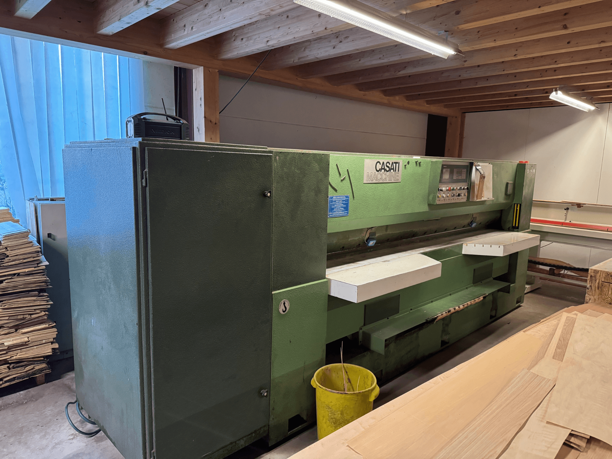 Máquina para trabajar la madera CASATI verde en un taller, vista frontal, con panel de control y superficies de trabajo blancas, rodeada de pilas de madera.