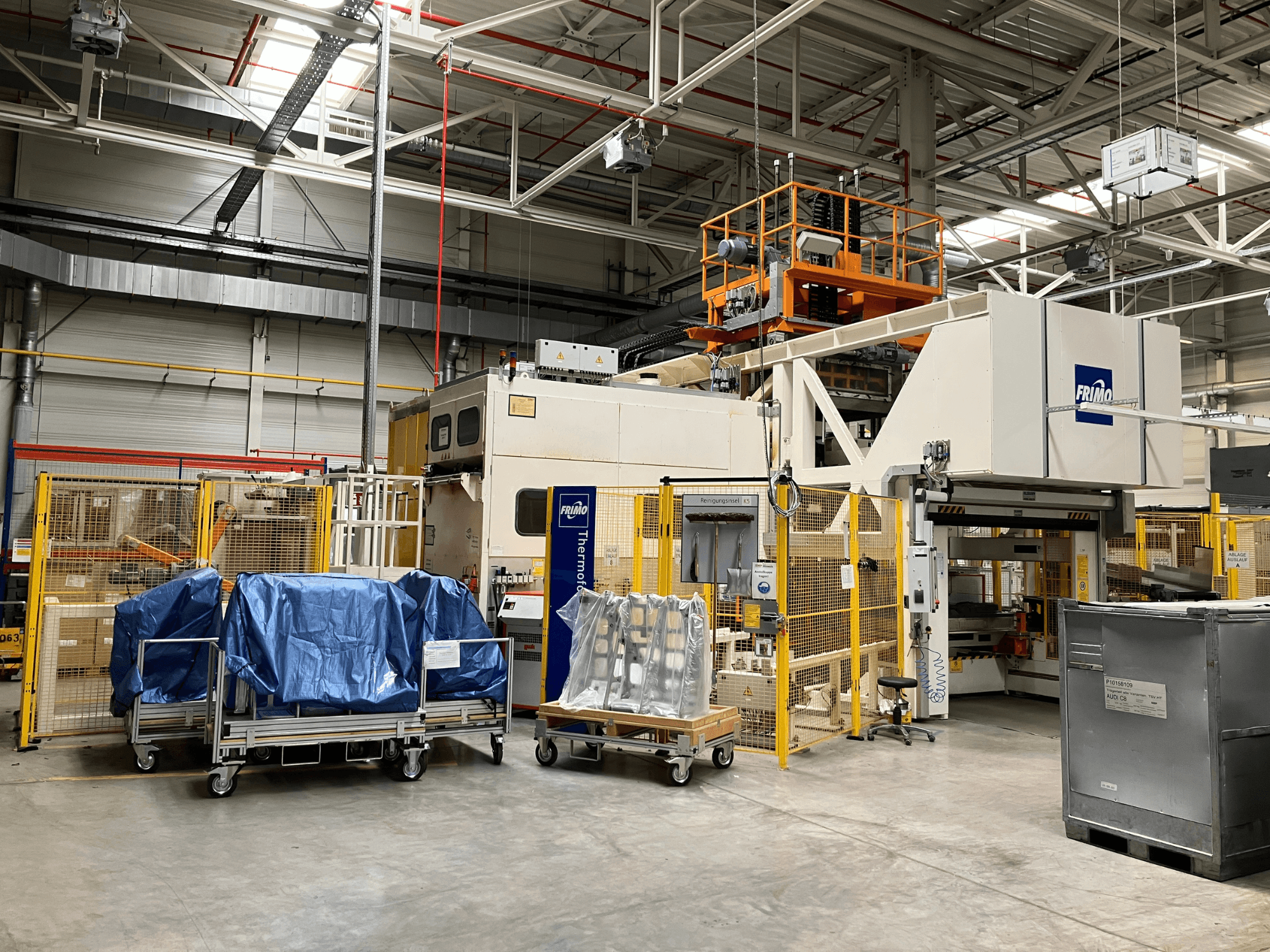 Máquina de procesamiento de plástico FRIMO FTL 80/200 Fullflex en una fábrica, vista frontal con lonas azules y barreras de seguridad visibles.