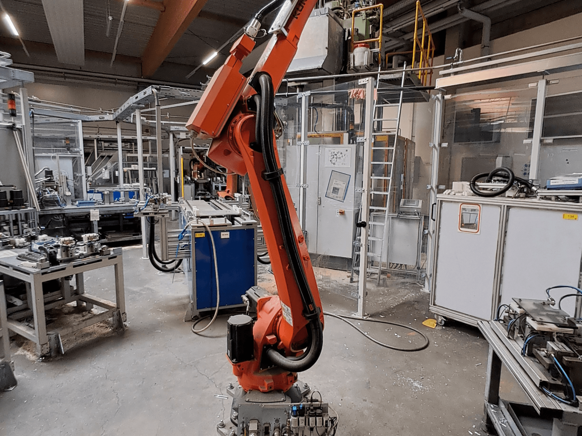Brazo robótico KUKA KR 26 R3100/SEL en un entorno industrial, visto desde un perfil lateral con equipos operativos al fondo.