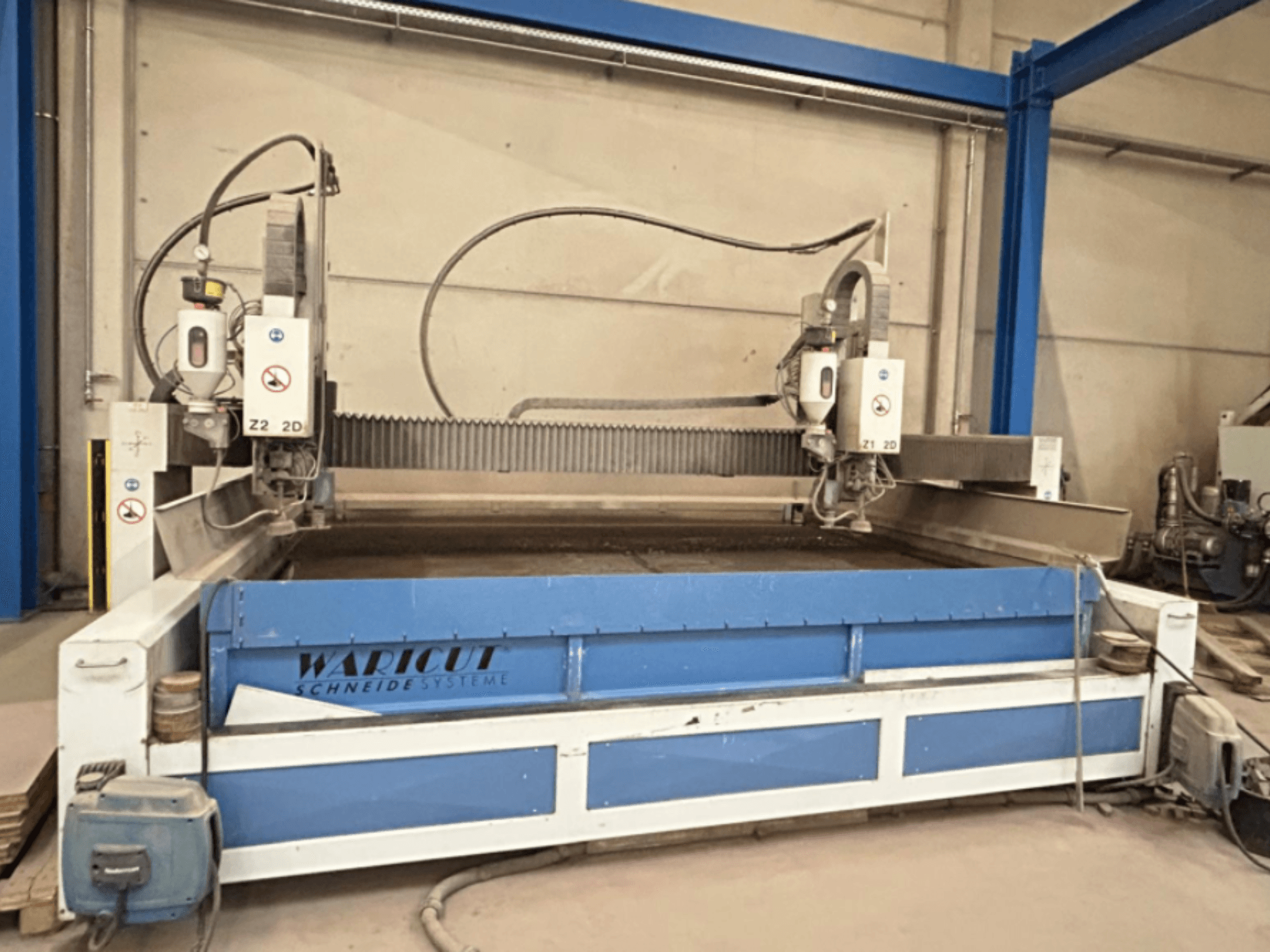 Máquina de corte por chorro de agua de Waricut, vista frontal; cuenta con dos cabezales de corte y una superficie de trabajo azul en un taller.