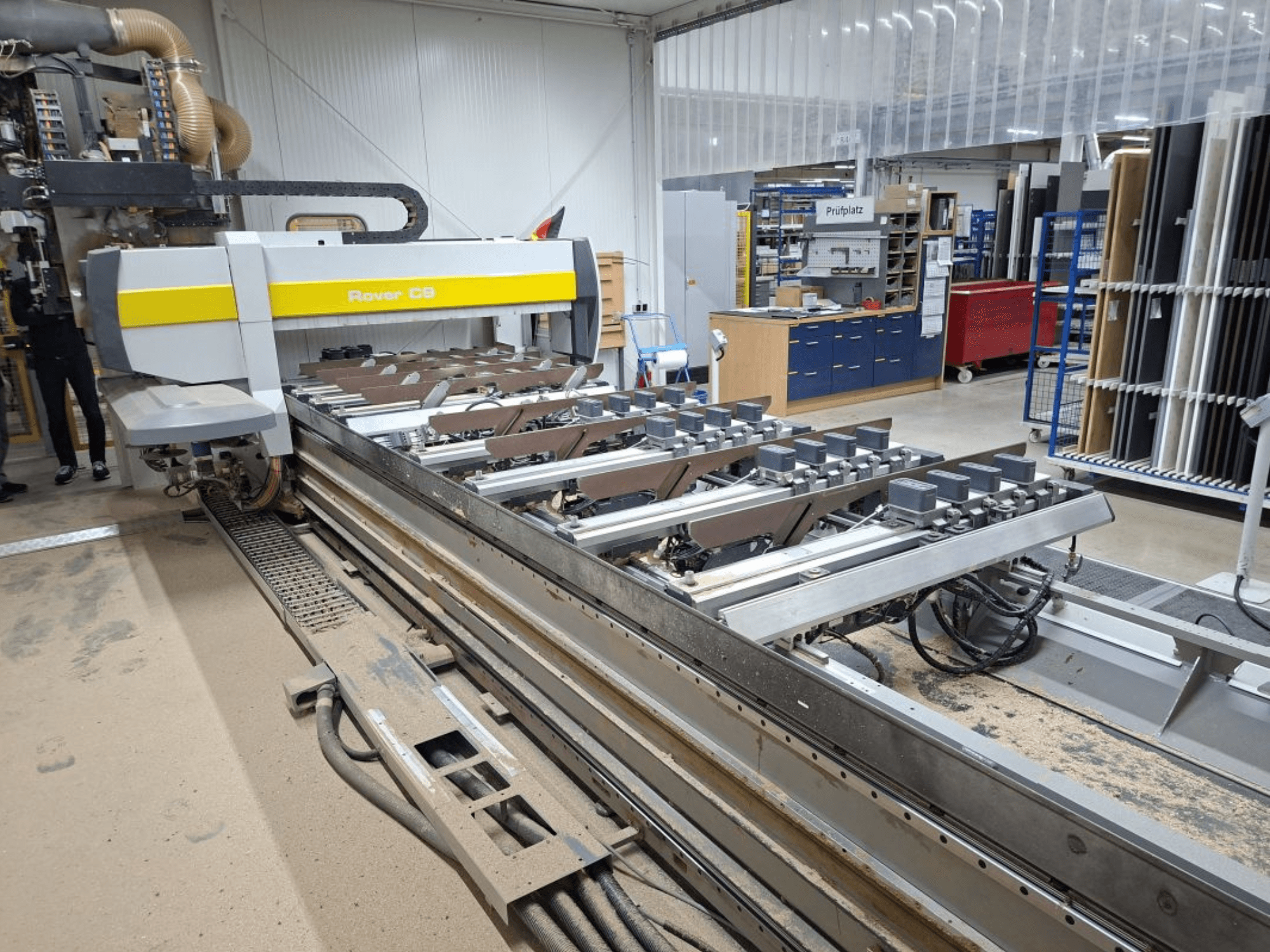 Máquina CNC Rover C9 en un taller; vista frontal que muestra las herramientas de corte y el sistema de transporte, con acumulación de polvo en el suelo.