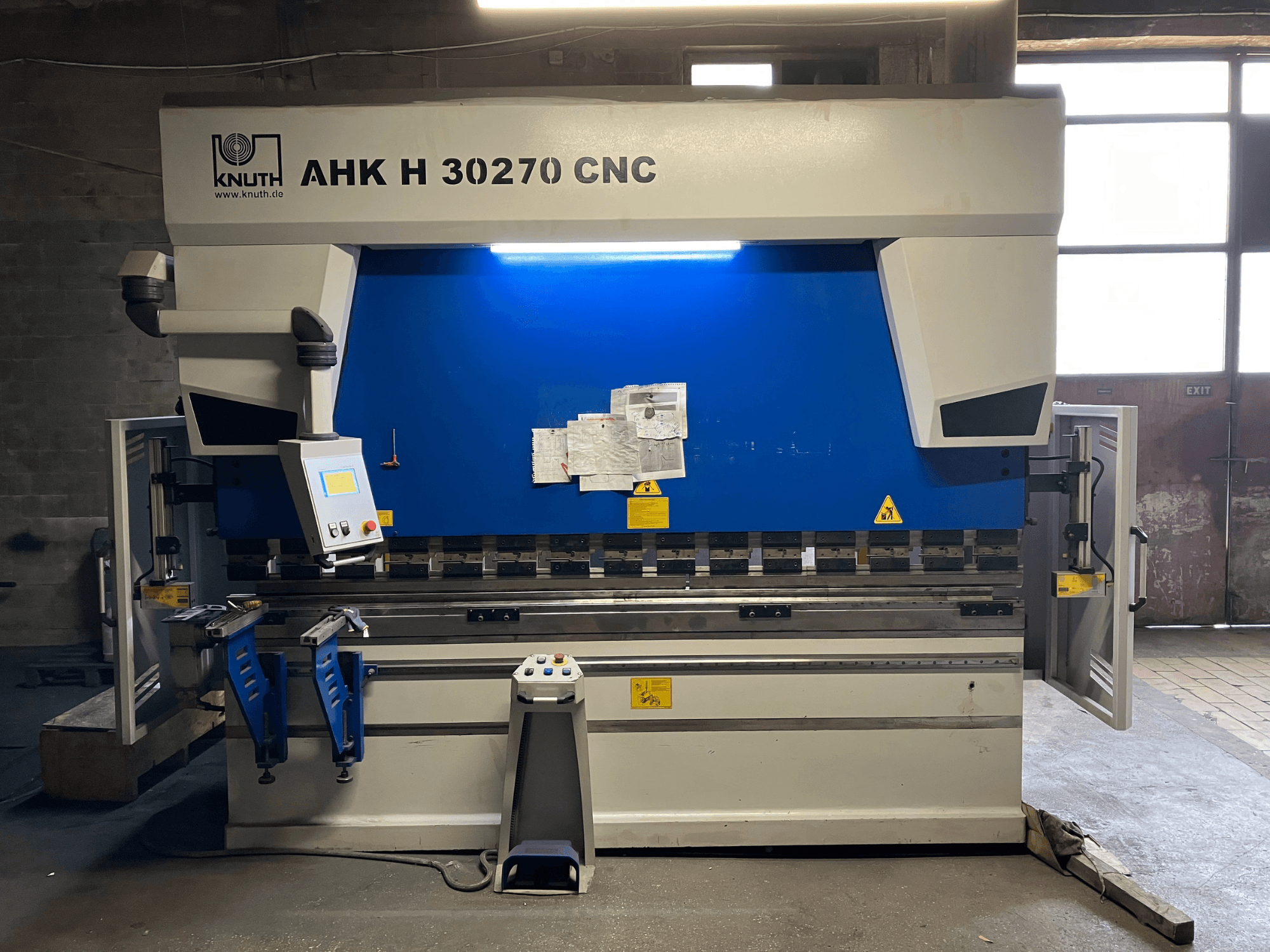 Plegadora CNC KNUTH AHK H 30270 en un taller, vista frontal, en la que se aprecian el panel de control y el diseño en azul y blanco, con la puerta lateral abierta.