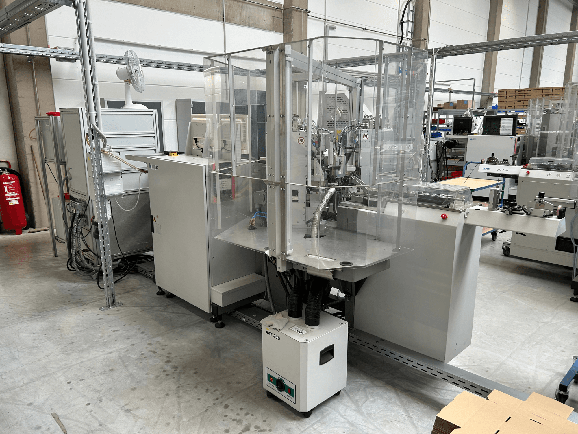 Equipo de automatización KOMAX Alpha 433 L en un entorno industrial. Cuenta con una cabina de seguridad transparente y varios paneles de control.