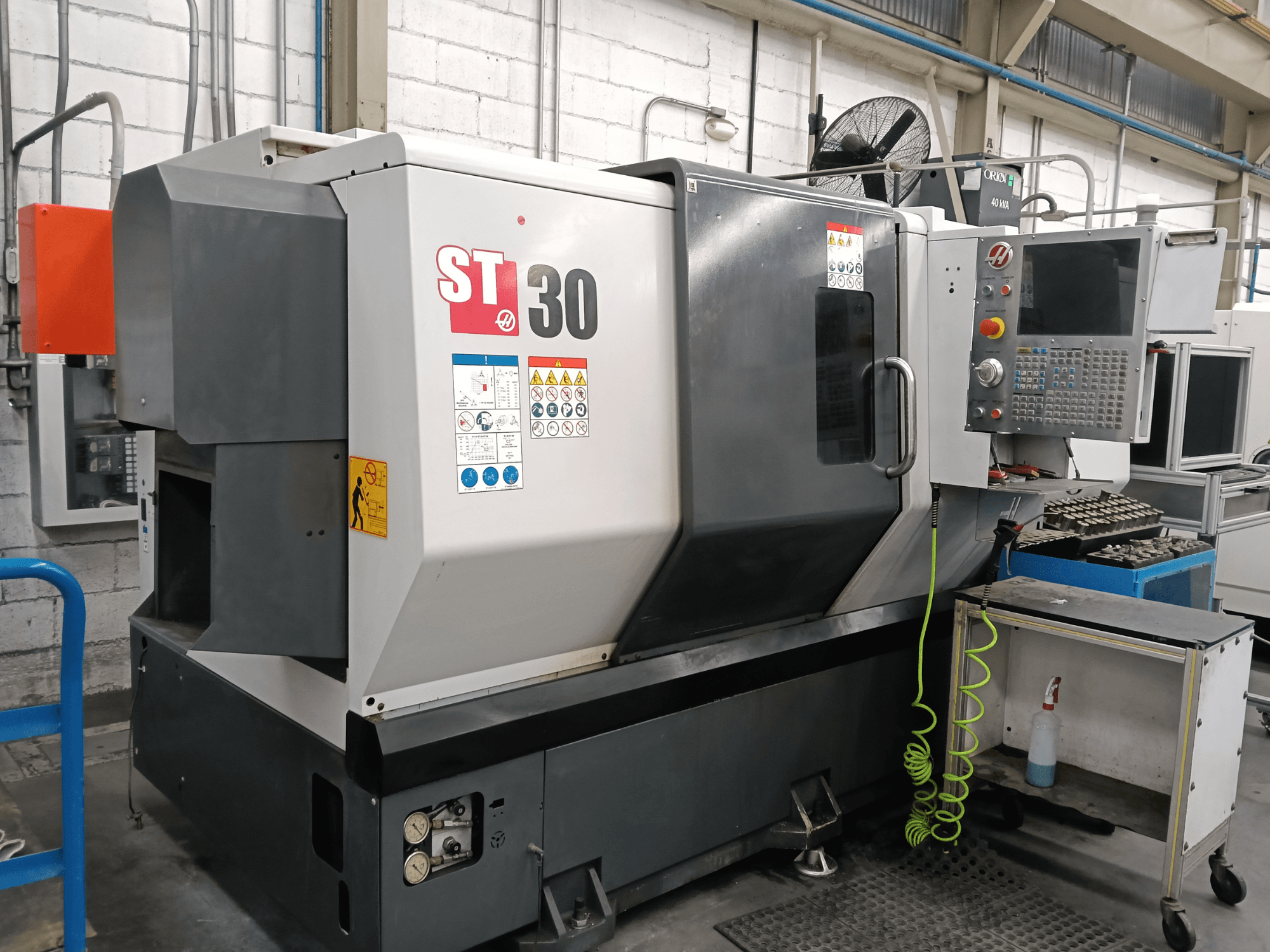 Texto alternativo: Vista frontal del torno horizontal HAAS ST-30 con el panel de control, las etiquetas de seguridad y las herramientas en un carro cercano.