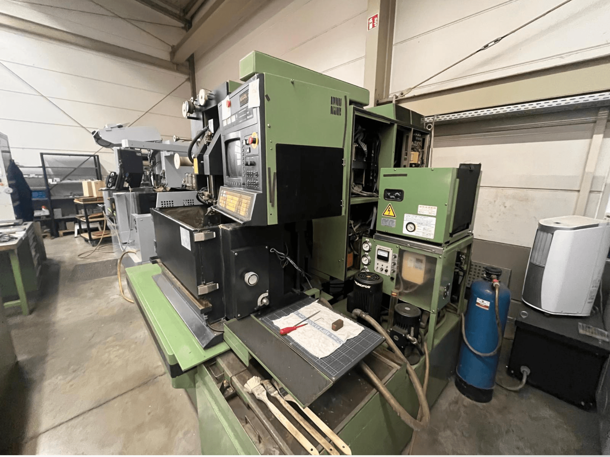 Máquina industrial verde con panel de control, vista lateral; incluye componentes hidráulicos y superficie de trabajo con herramientas.